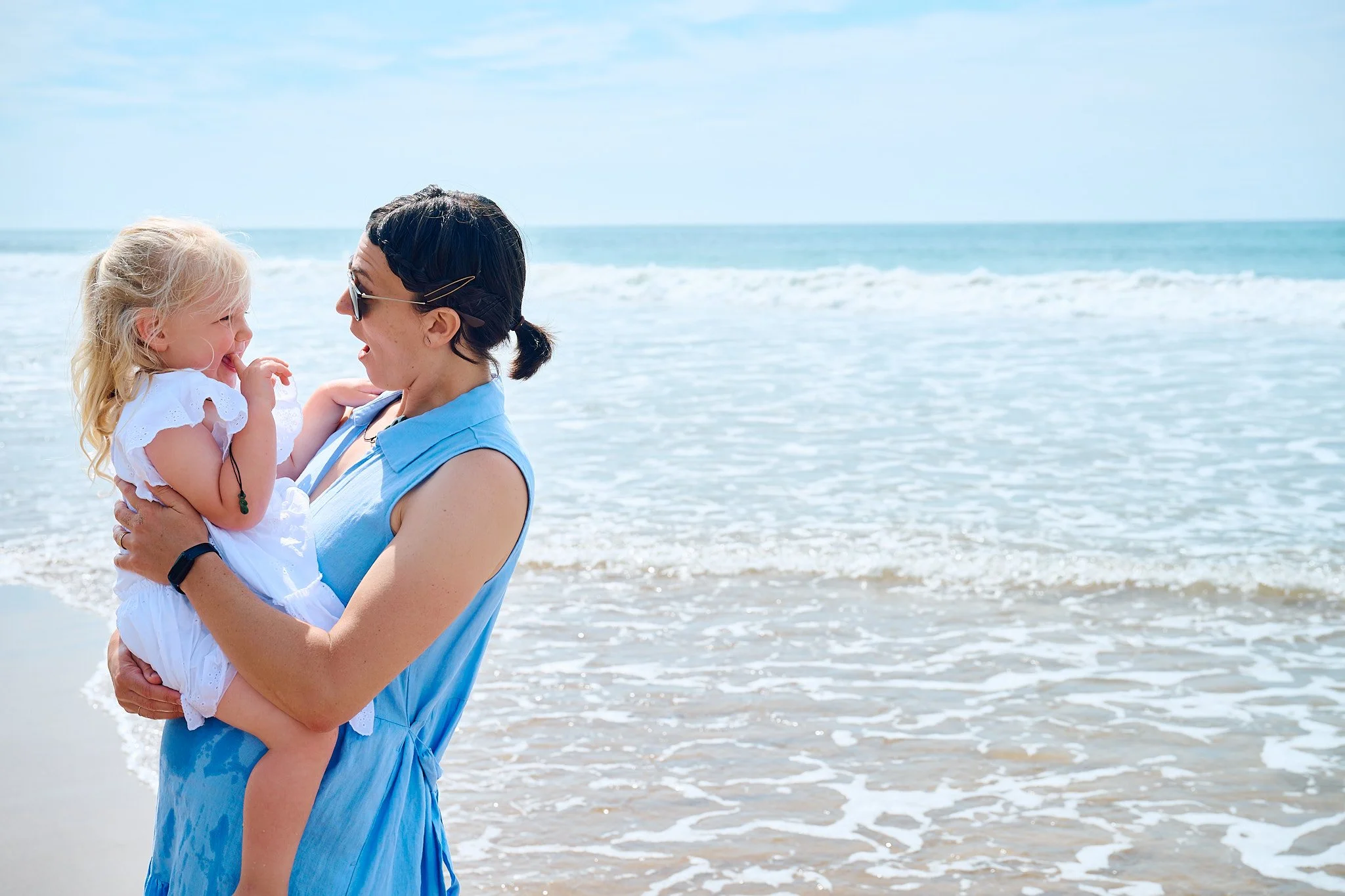 Woman holding a child laughing together at the beach, with ocean waves in the background.