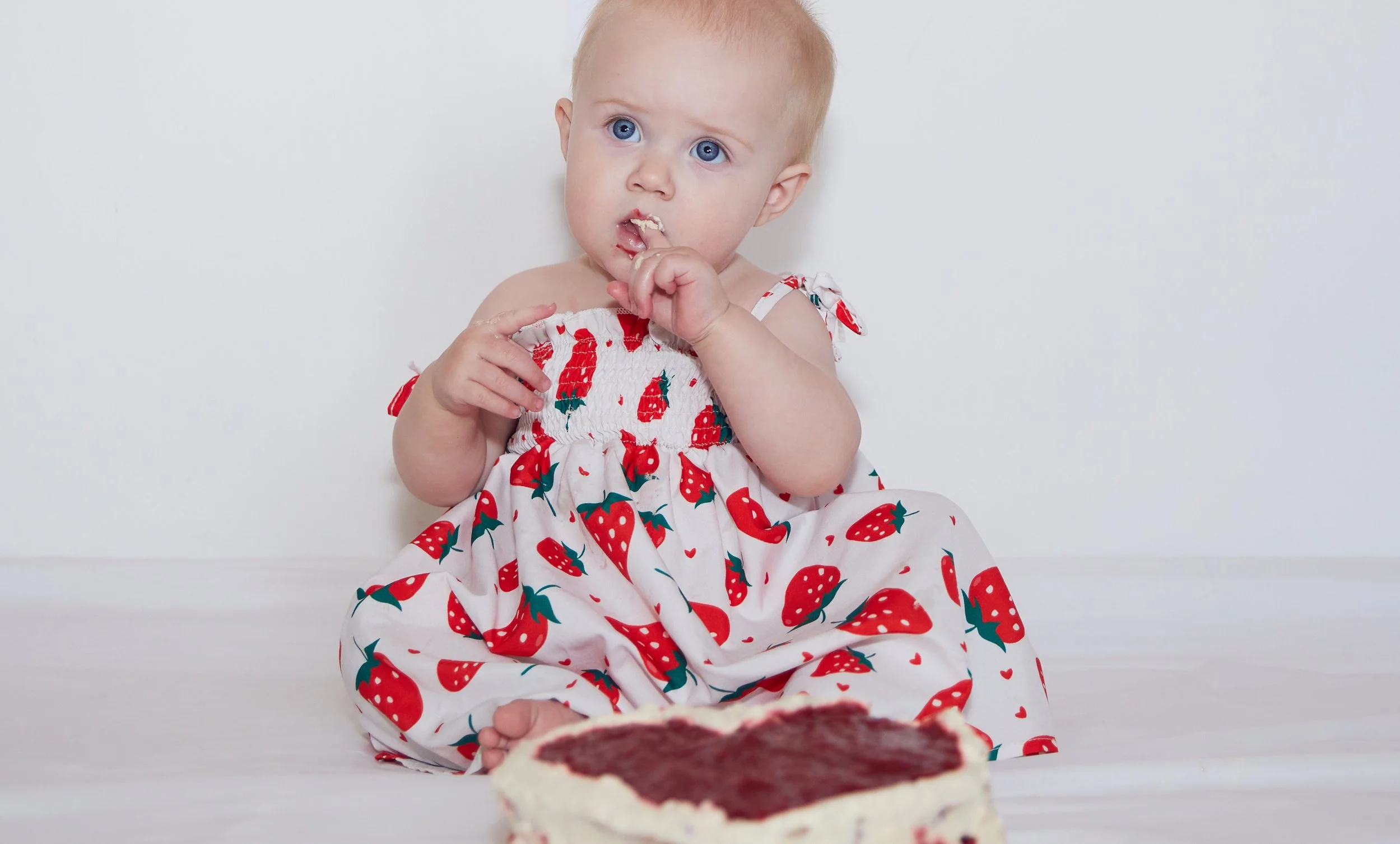 Baby in strawberry-patterned dress sitting with a cake, touching its face with frosting.