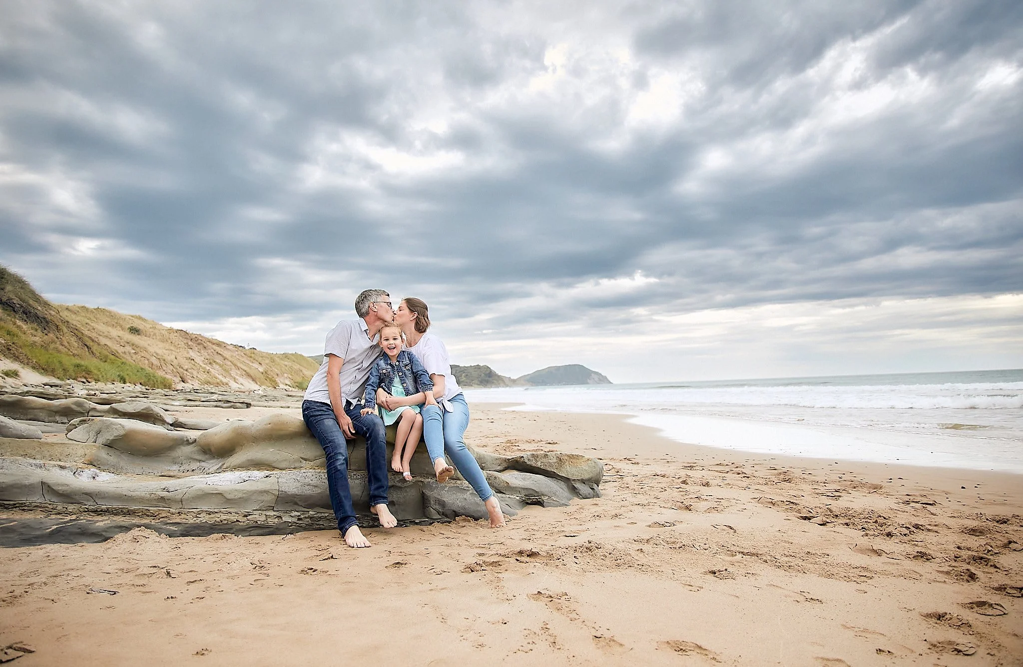 Family sitting on a beach with rocky shore, cloudy sky, ocean in the background.