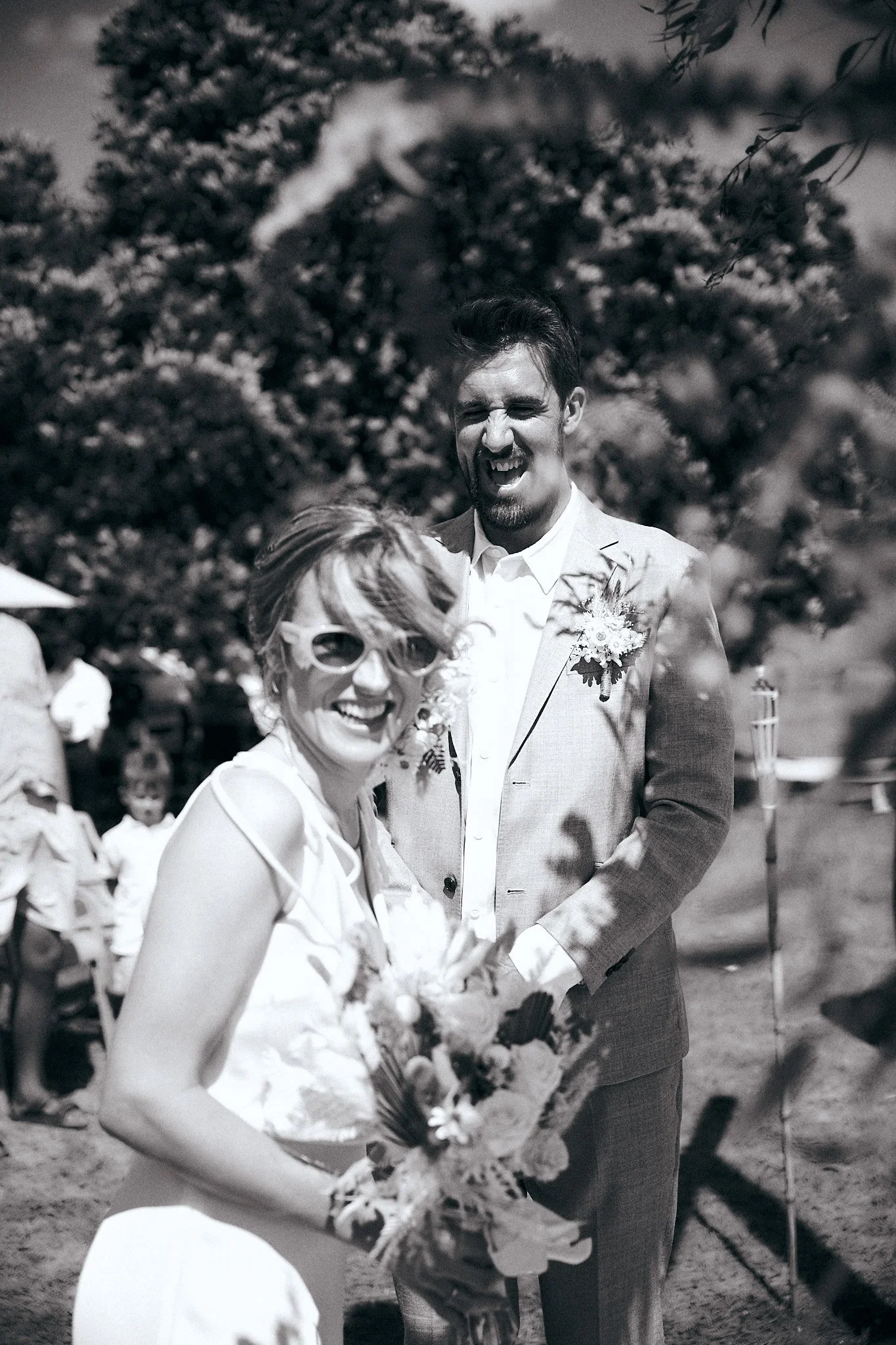 Black and white photo of a smiling bride holding a bouquet and a groom standing behind her, outdoors.