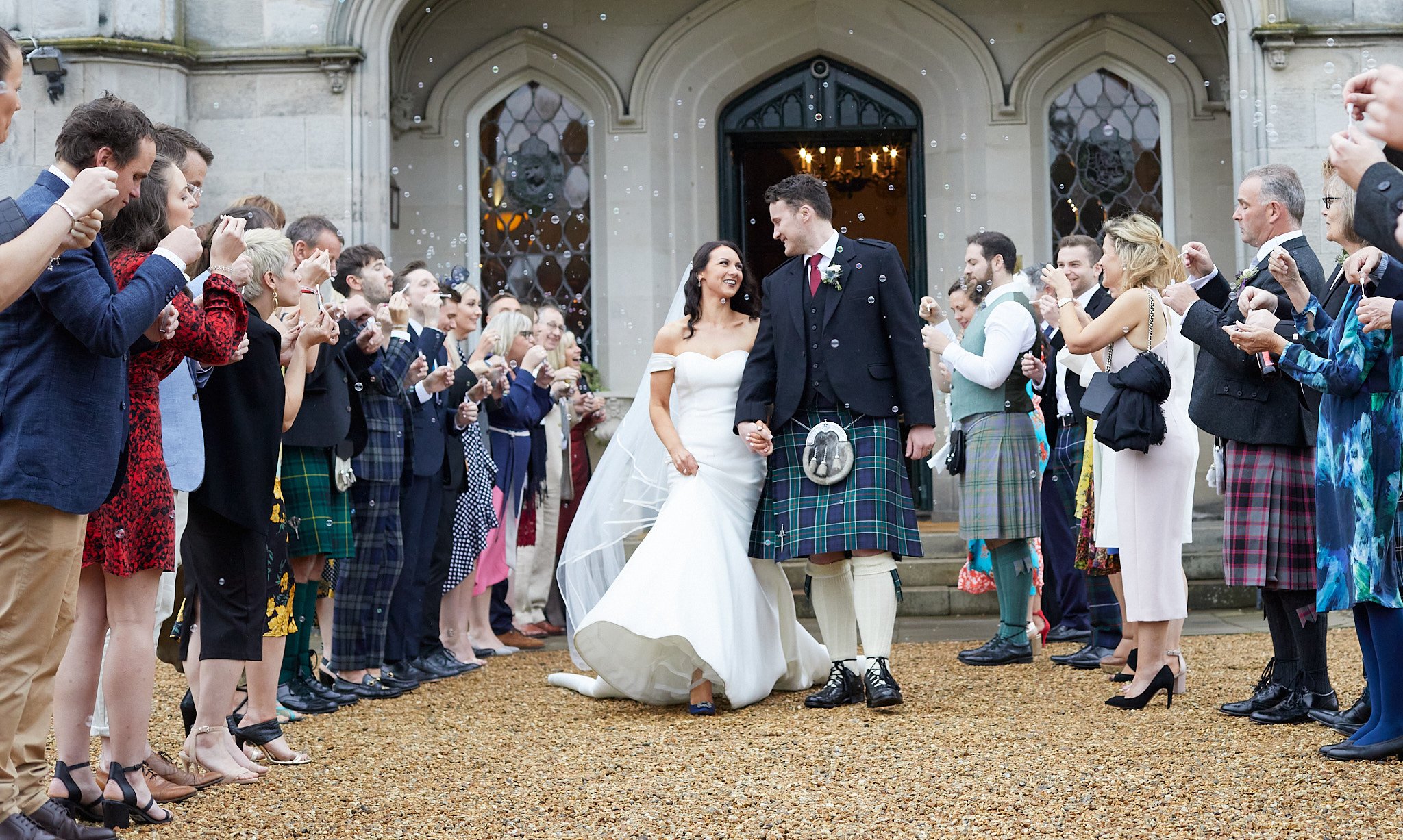 Bride and groom in traditional Scottish attire walking through a crowd of wedding guests outside a stone building.