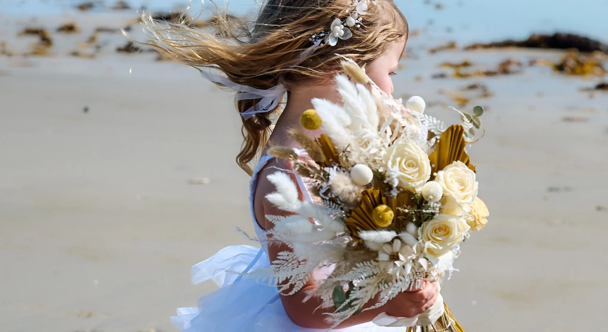 Young girl in white dress holding a bouquet of flowers on a beach.