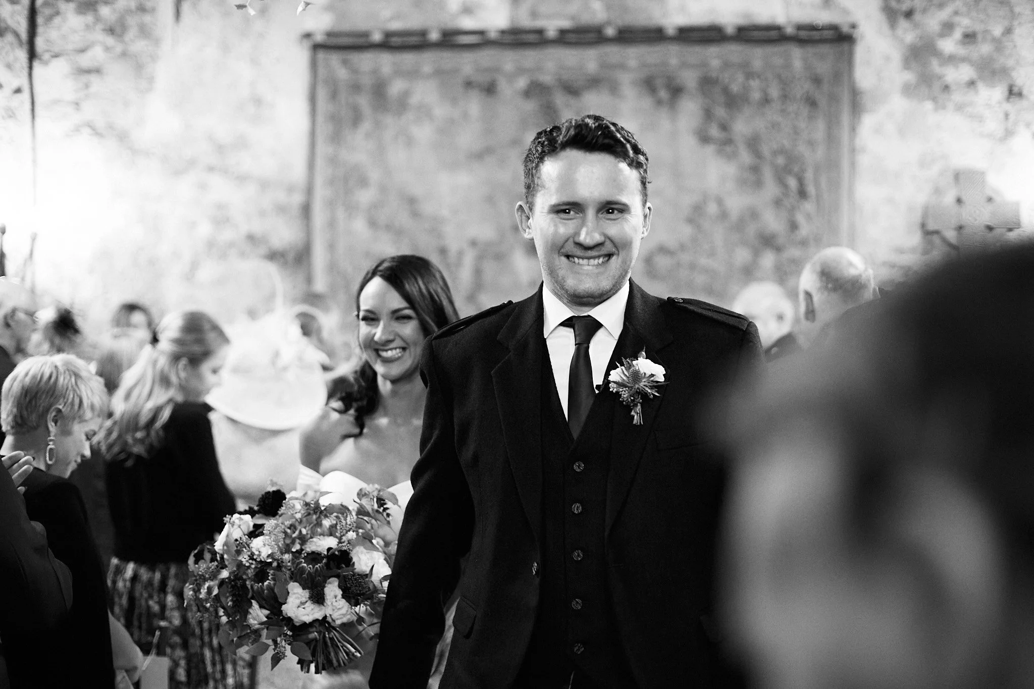 Bride and groom smiling at a wedding ceremony, with the bride holding a bouquet and guests in the background. Black and white photograph.
