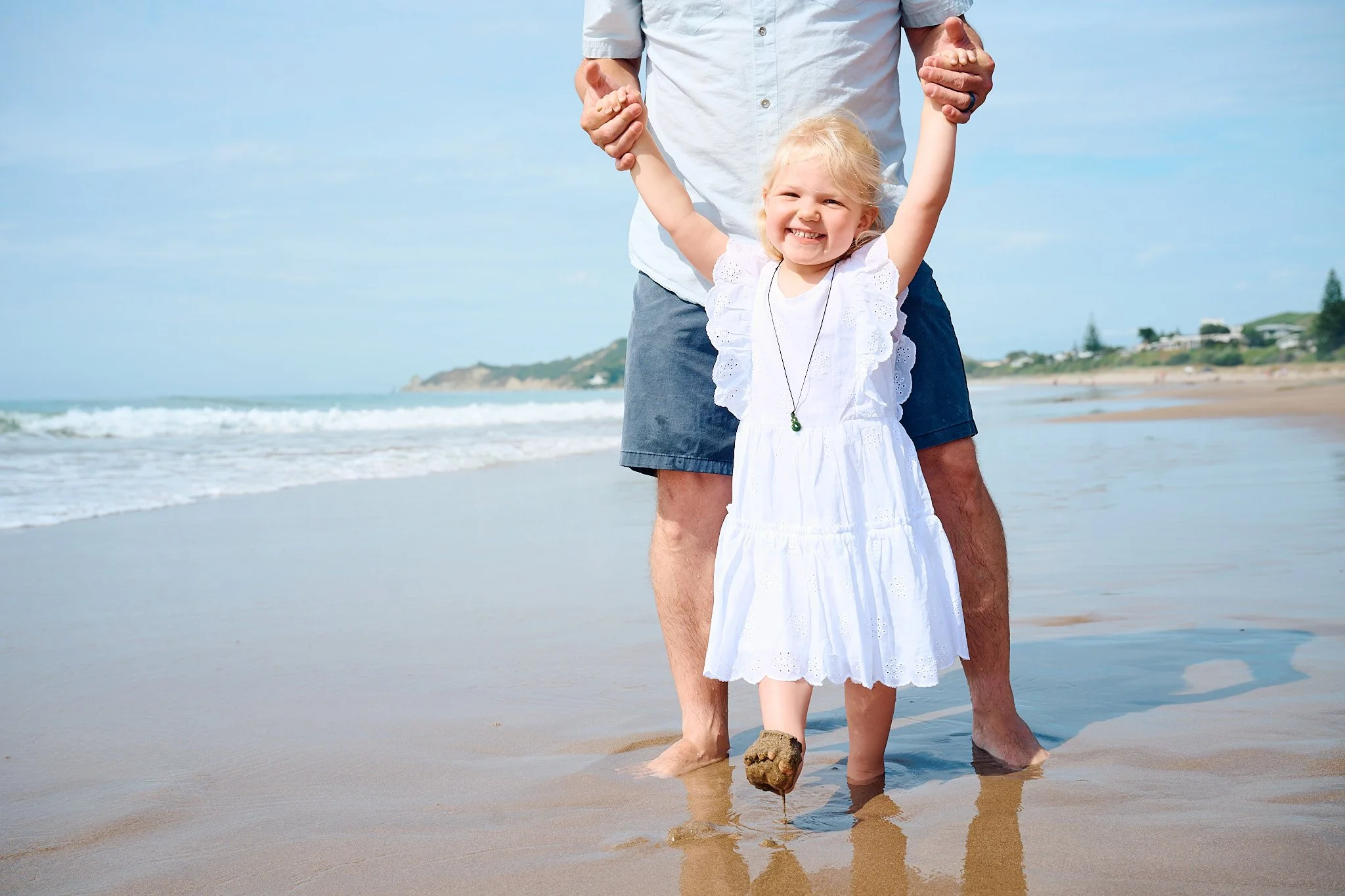 Child in white dress smiling while being held by an adult at the beach.