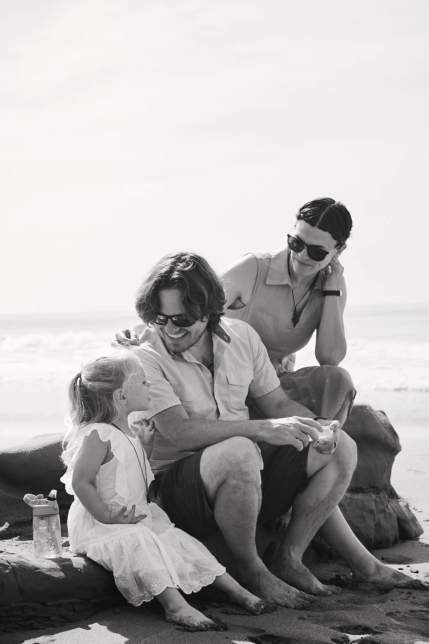 Black and white photo of a family sitting on a beach, including a man in sunglasses interacting with a young girl in a dress, and a woman wearing sunglasses nearby.