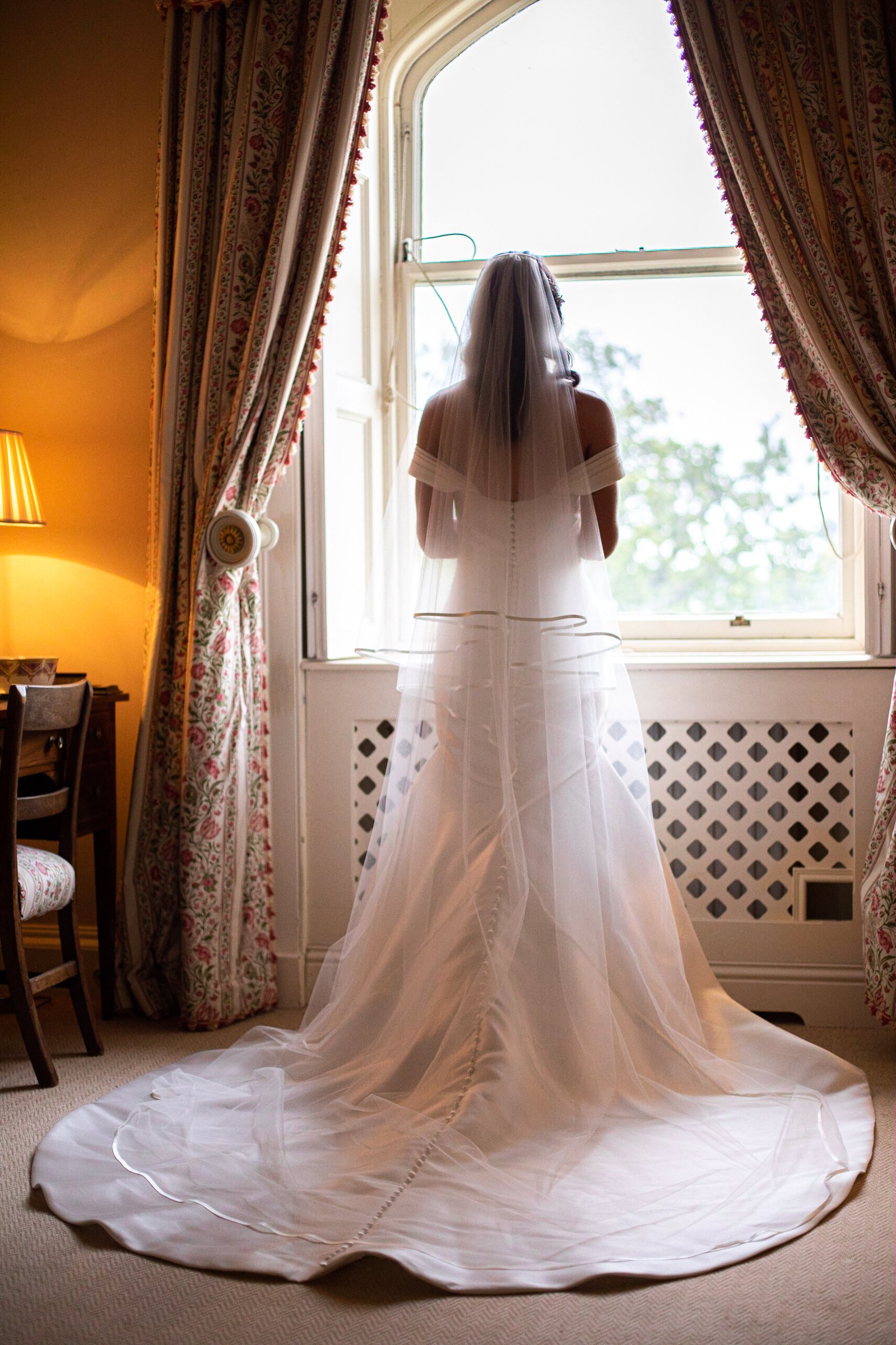 Bride in white dress and veil standing by a window, with elegant curtains and a side table with a lamp.