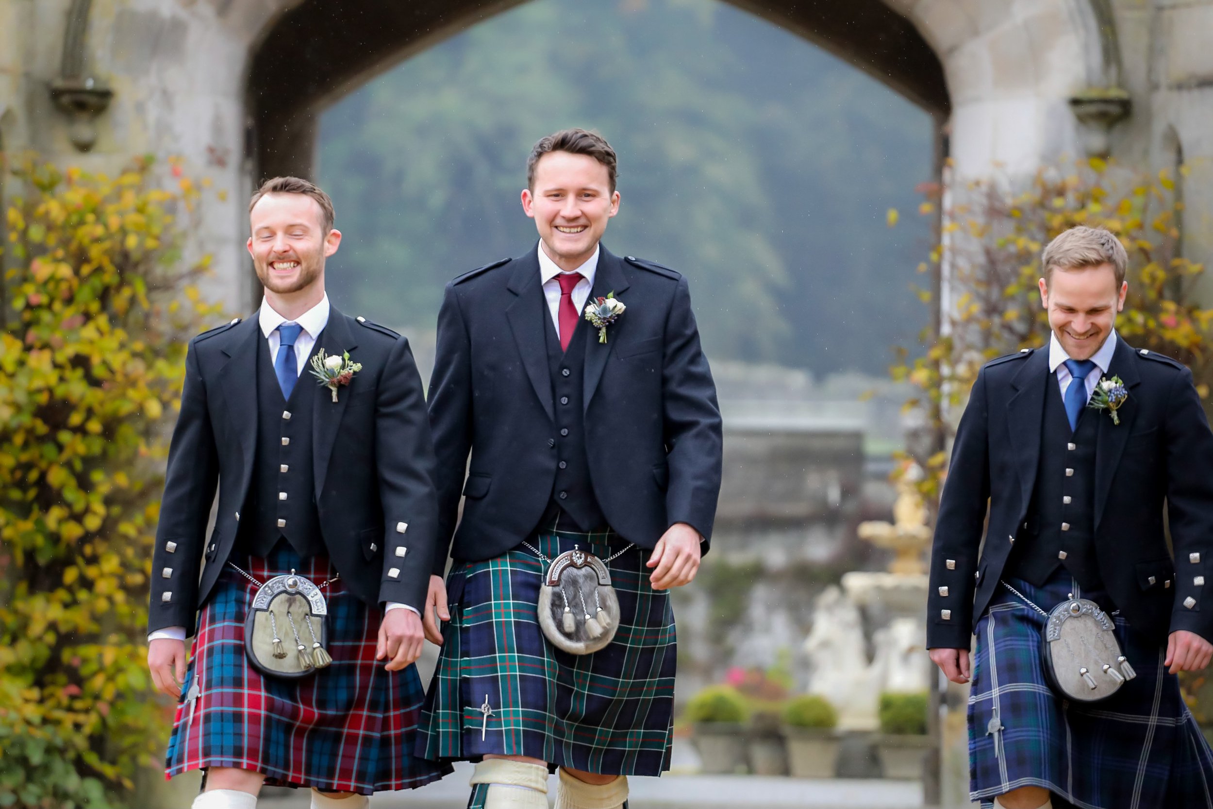 Three men wearing traditional Scottish kilts walking under an archway.