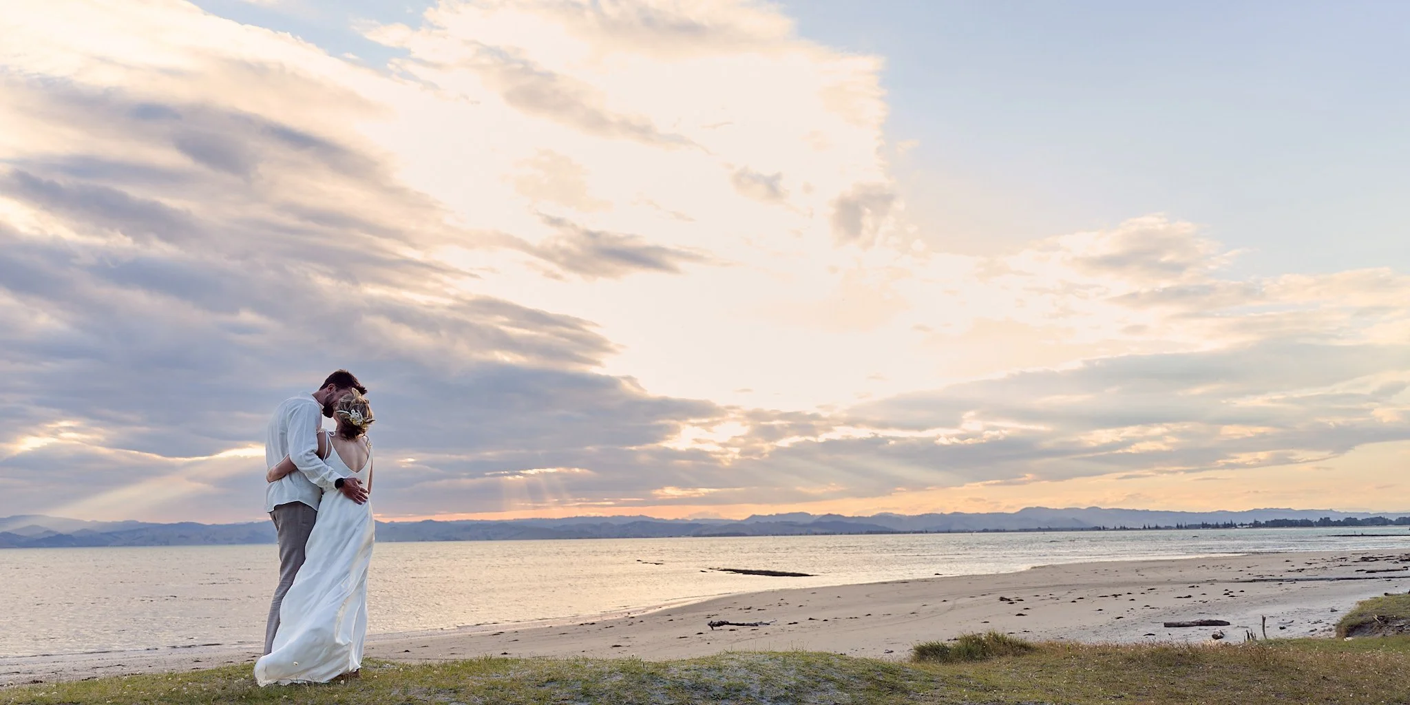Bride and groom embracing on a beach at sunset, with ocean and clouds in the background.