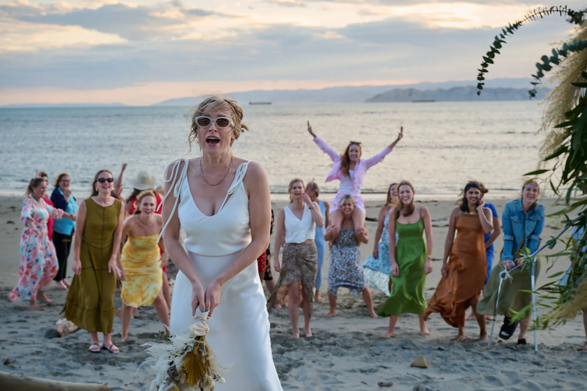 Bride in white dress preparing to throw bouquet on beach with excited group of women in dresses cheering behind her.
