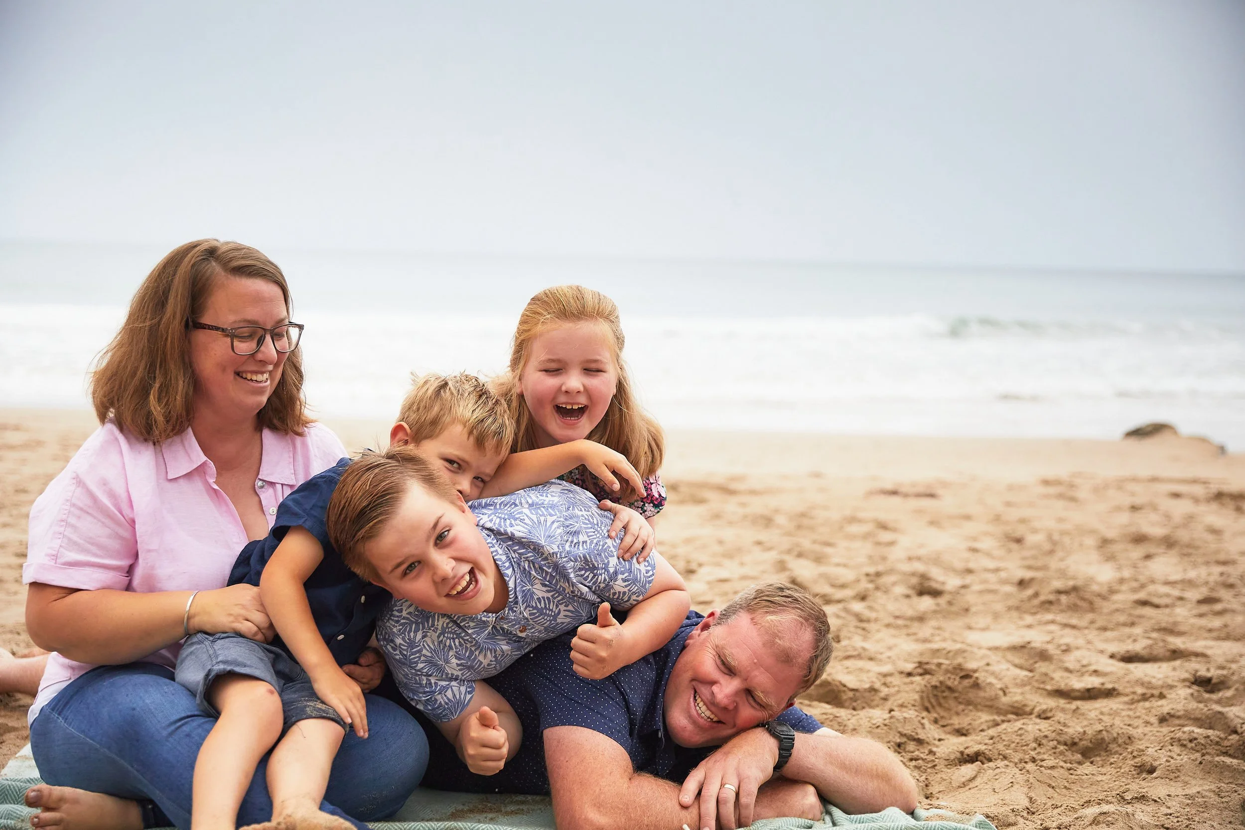 Family of five laughing on a beach, with children playful on top of father, mother sitting beside them. Sea and sand in the background.