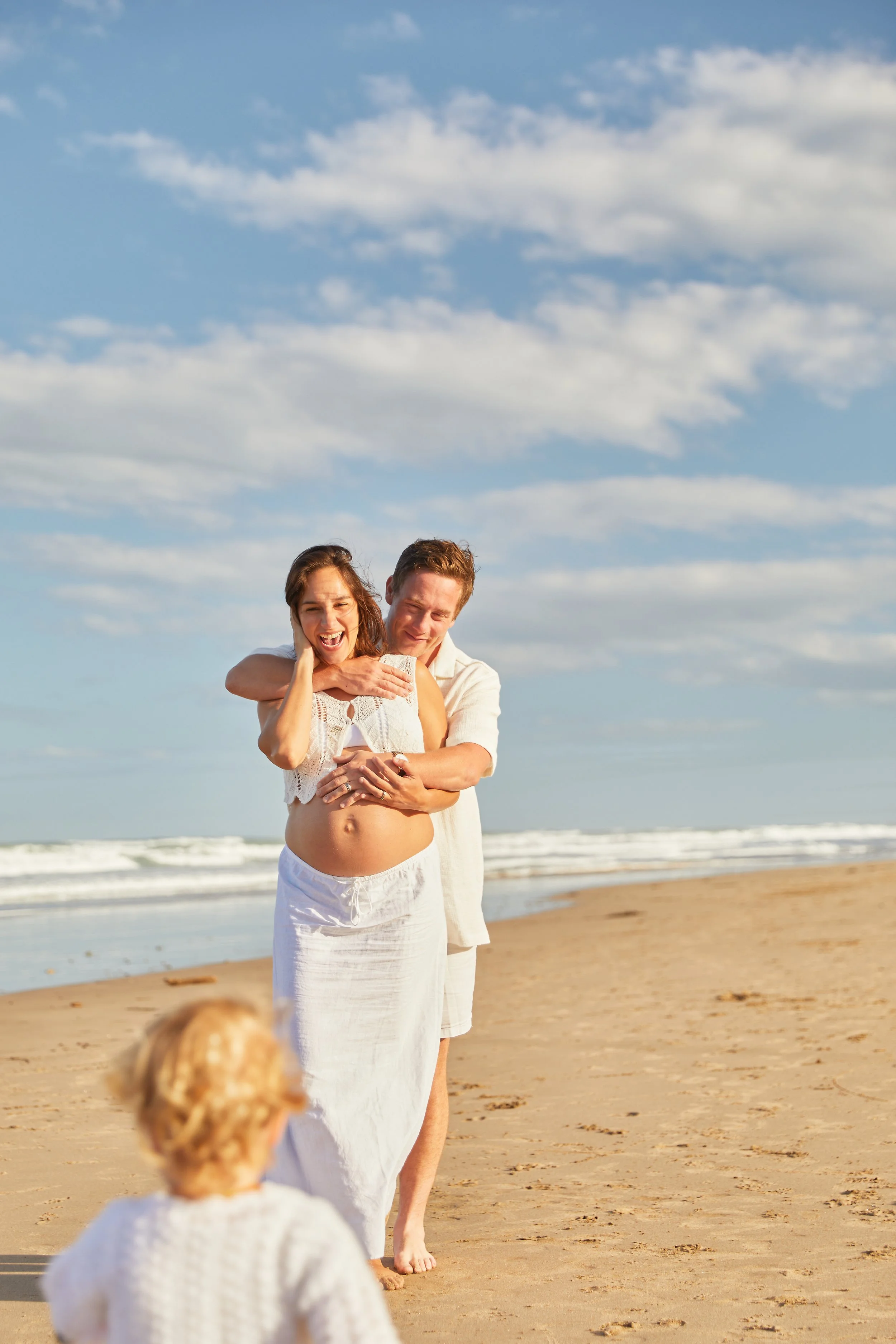 A pregnant woman with a man embracing her from behind on a beach, while a child stands in the foreground.