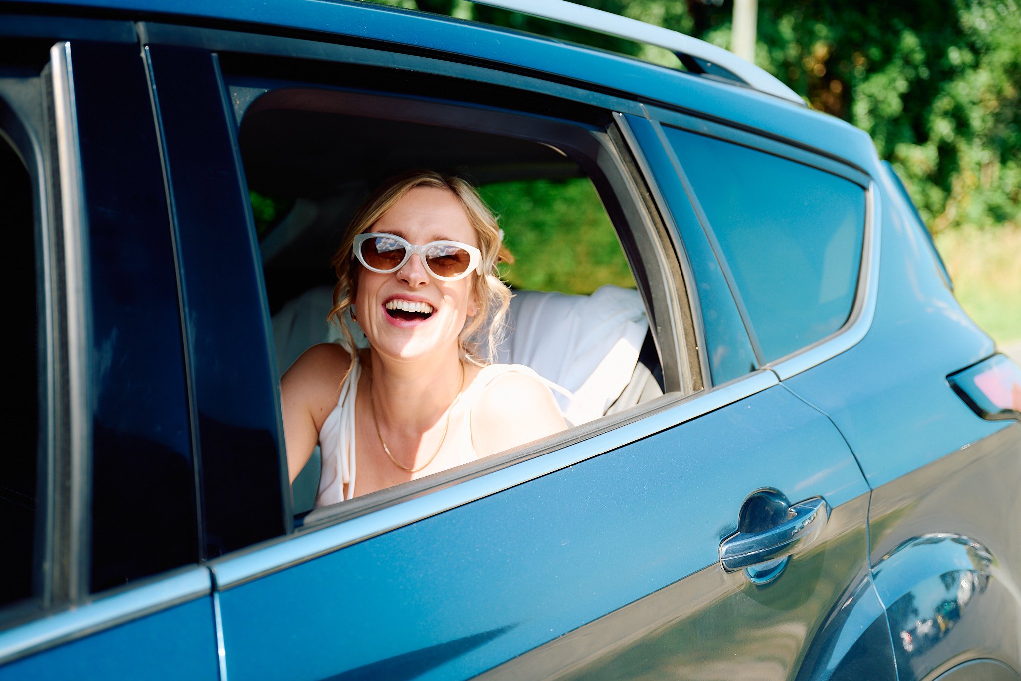 Woman wearing sunglasses leaning out of a car window, smiling.