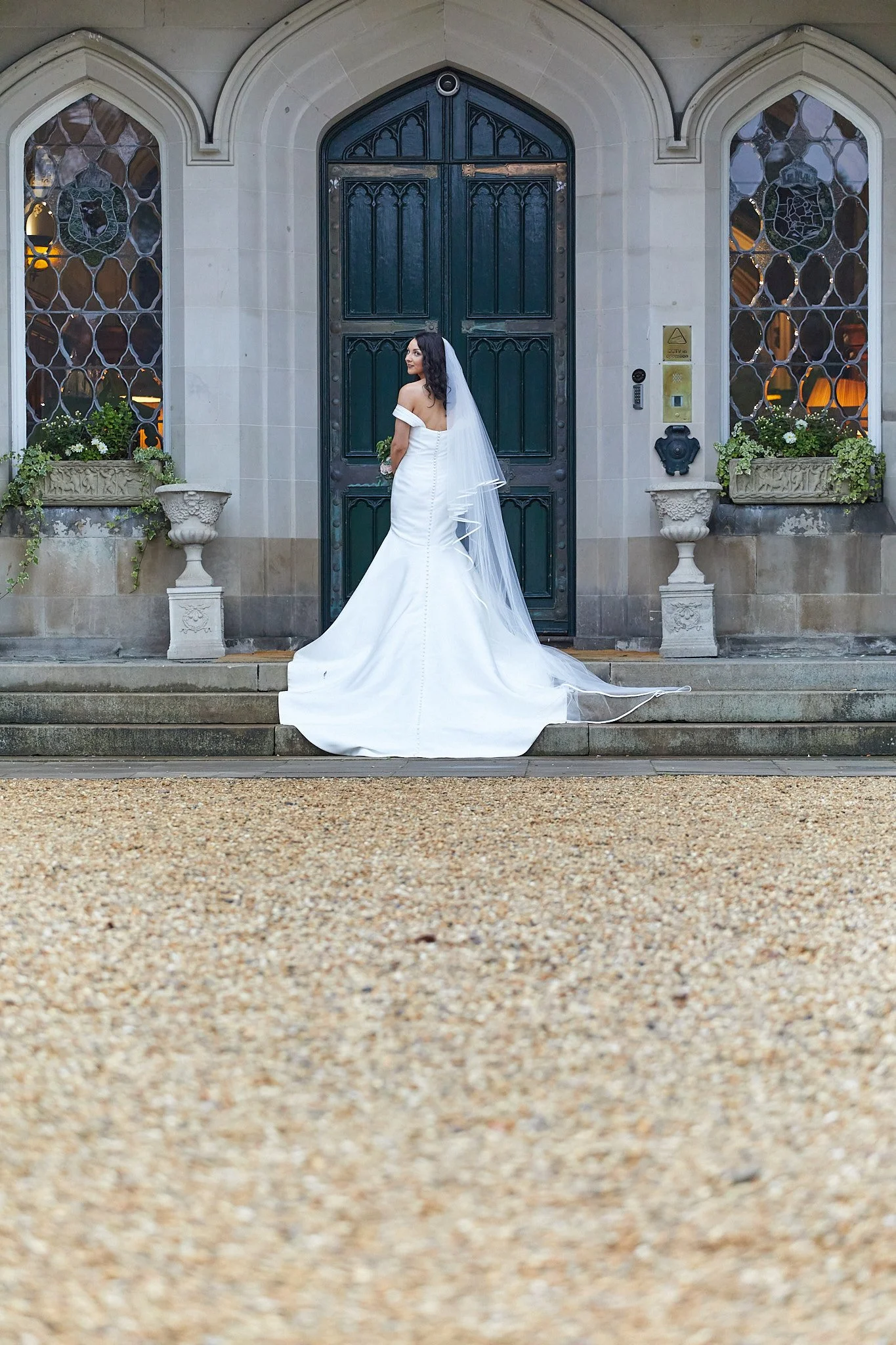 Bride in white gown and veil standing in front of ornate door and windows.