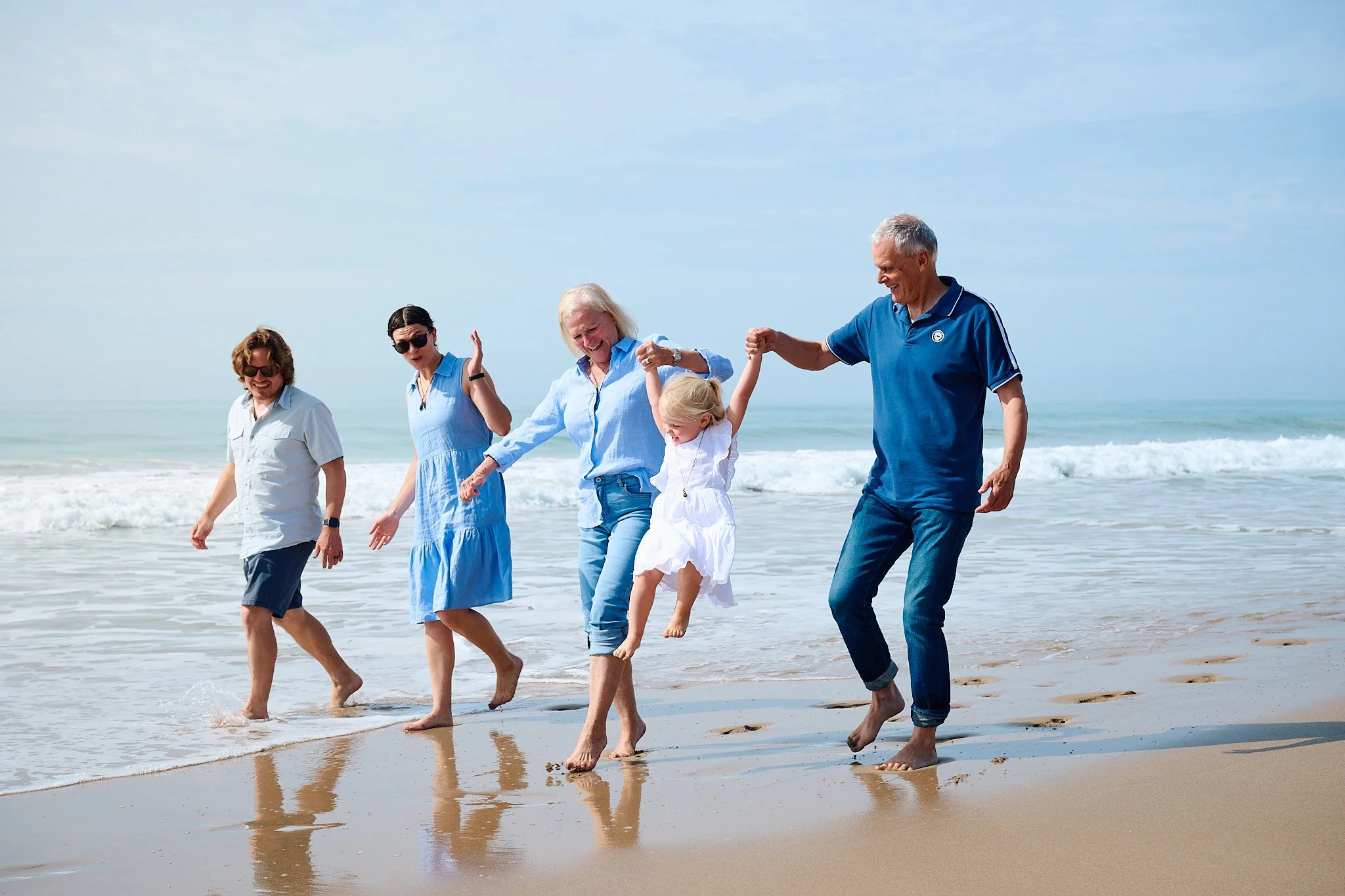 A family of five walking on the beach, including two older adults, two younger adults, and a child in the middle being swung in the air, with ocean waves in the background.