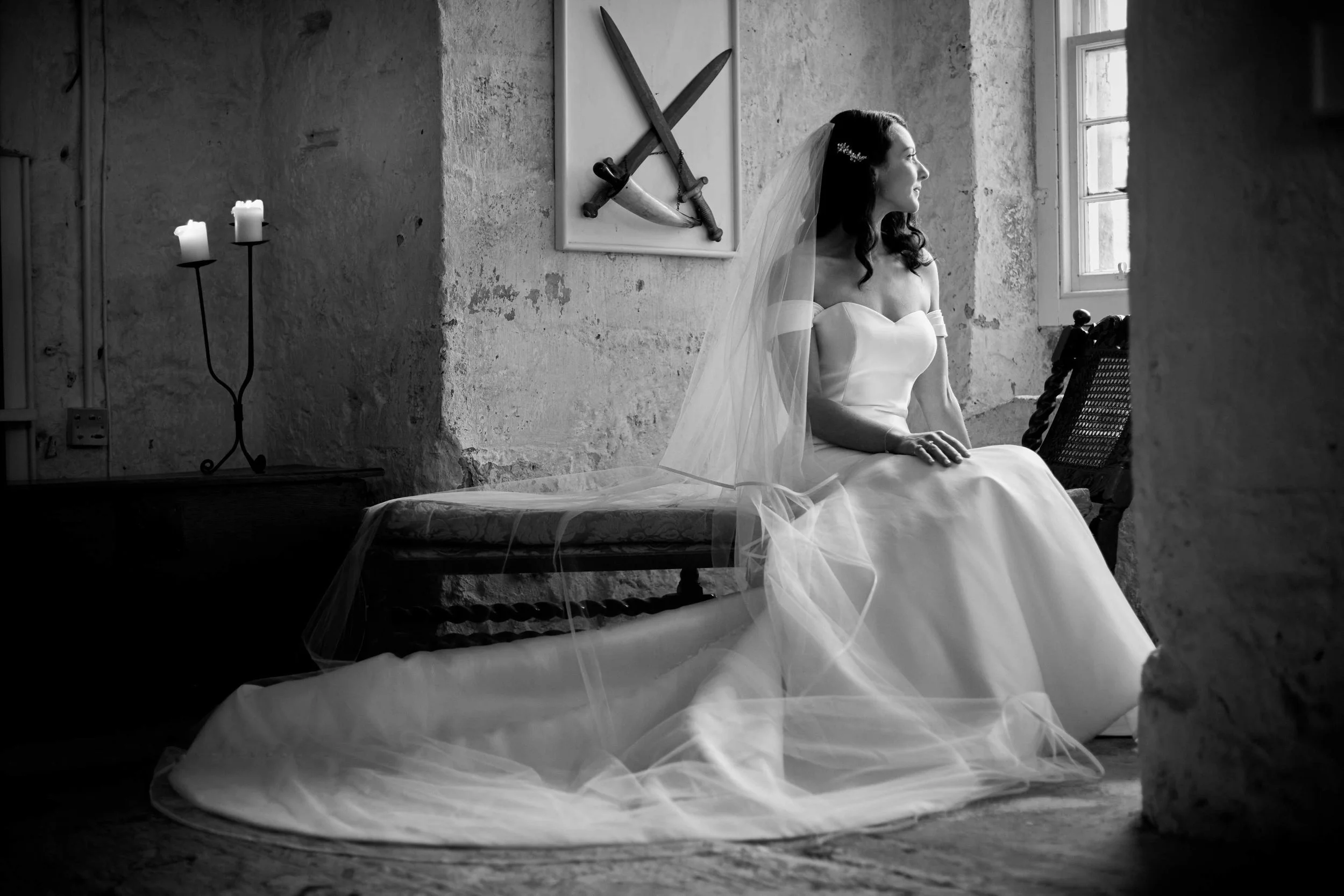 Bride in wedding dress sitting near window, old stone room, with candles and swords on wall, black and white photo.