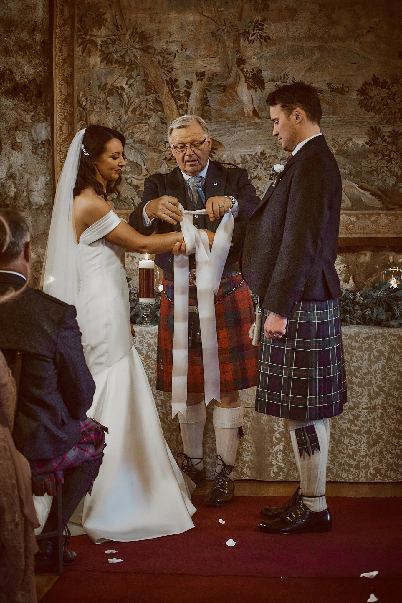 A wedding ceremony with a couple in traditional attire and officiant performing a handfasting ritual with ribbons.