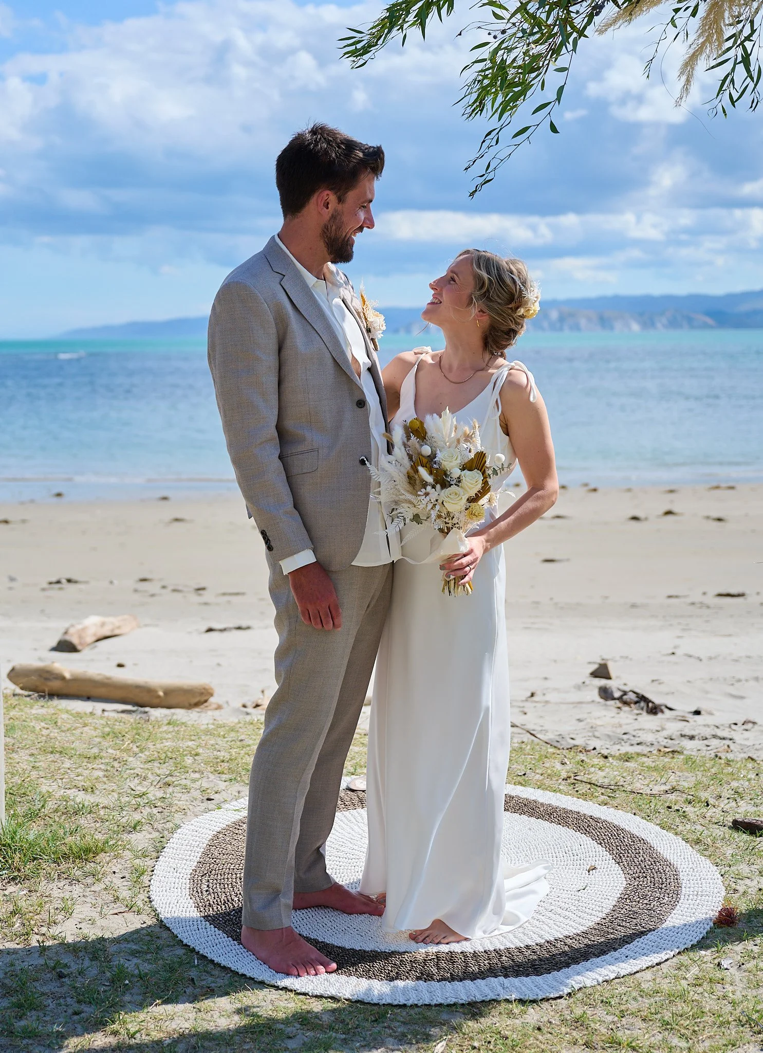 Bride and groom standing on beach, barefoot, smiling at each other, with a scenic ocean background.
