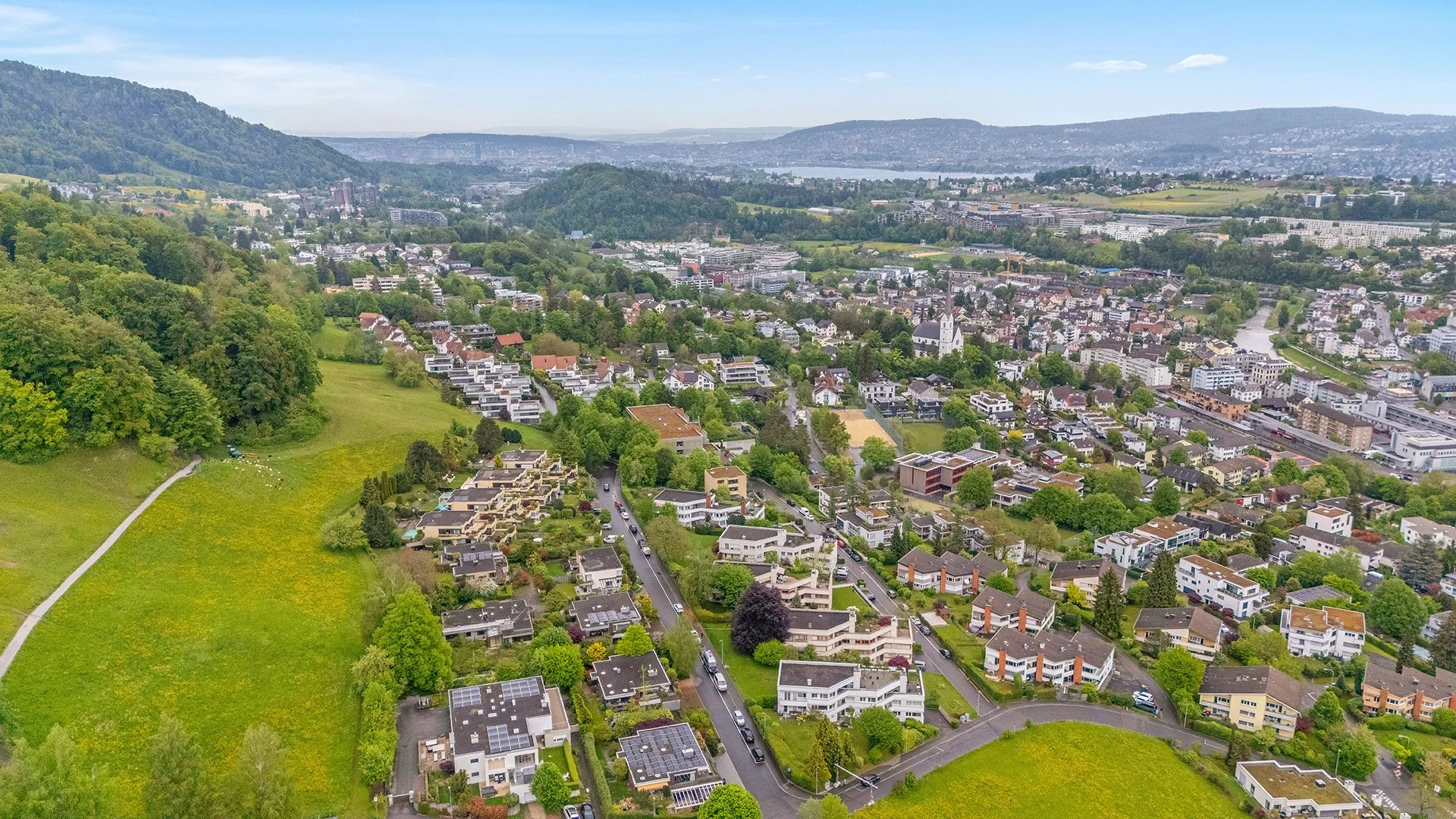 Luftaufnahme einer grünen Schweizer Stadt mit Wohnvierteln, Feldern, bewaldeten Hügeln und See im Hintergrund