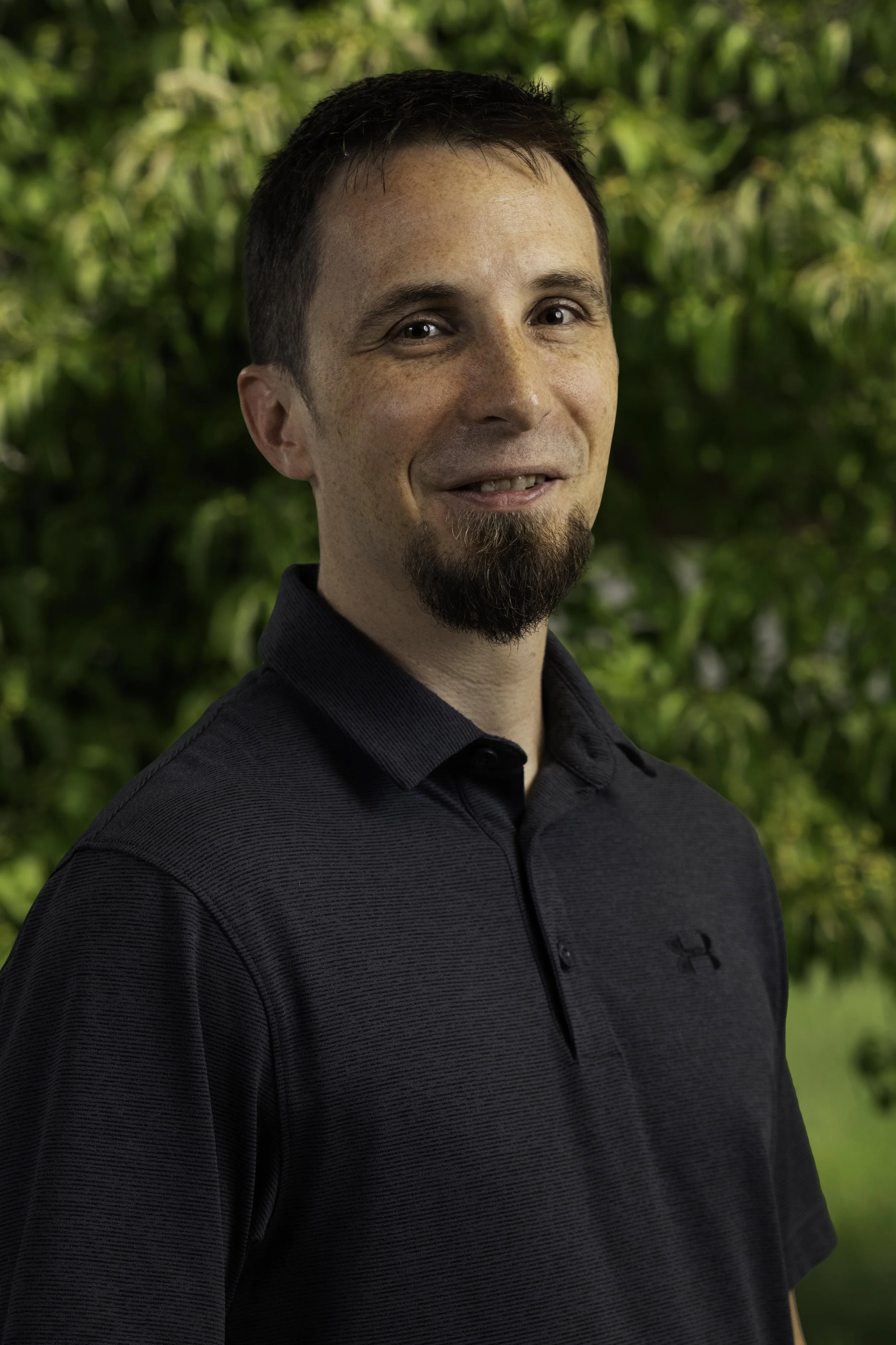 A man with short dark hair, a goatee, and wearing a beige button-up shirt standing outdoors in a park with trees and benches in the background during autumn.