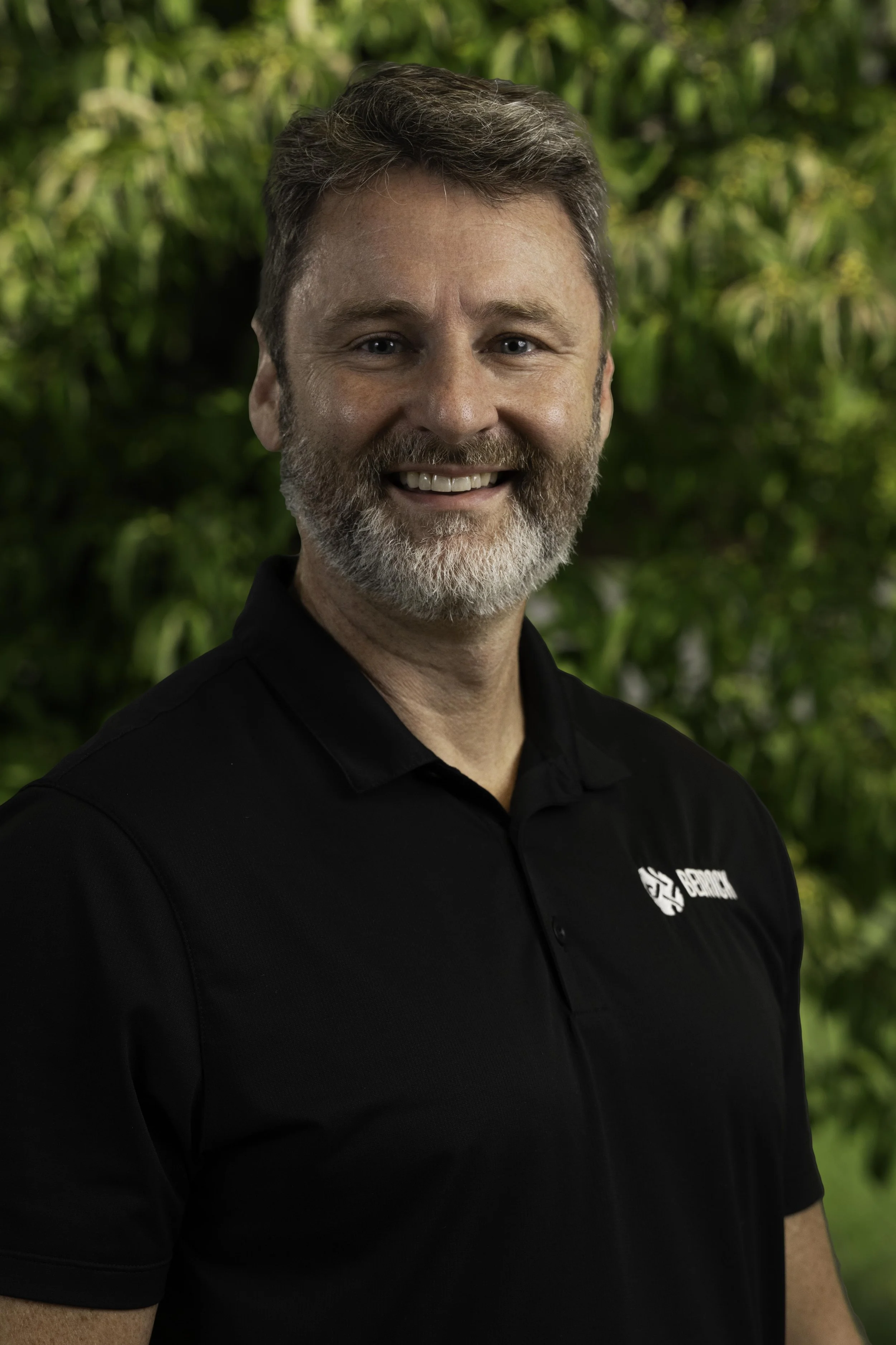 Smiling middle-aged man with a beard in a gray hoodie outdoors on a sunny autumn day, standing in front of trees with yellow leaves and a park path.