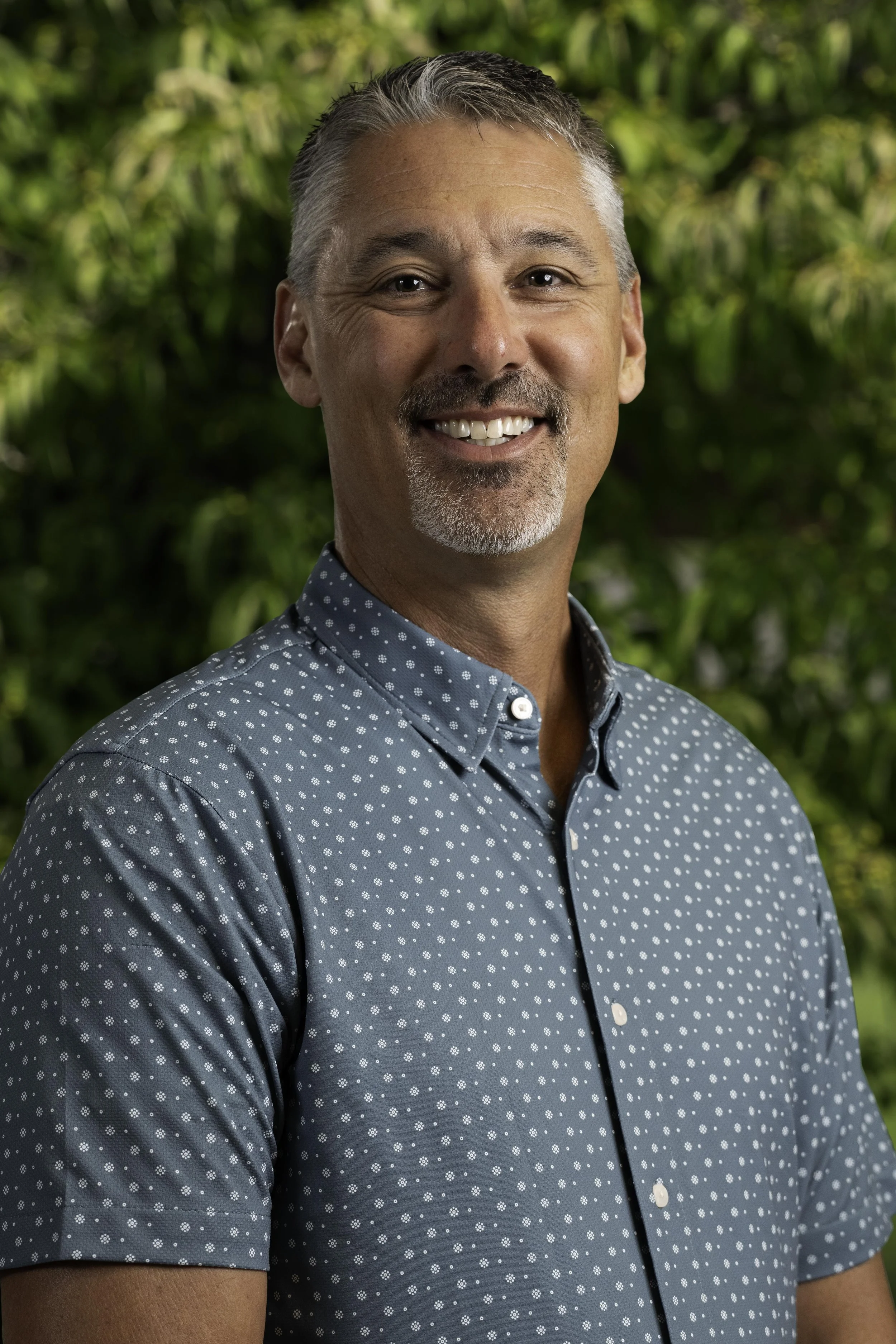 Smiling middle-aged man with gray hair and beard, wearing a red plaid shirt, outdoors in a park during fall with yellow and brown leaves in the background.