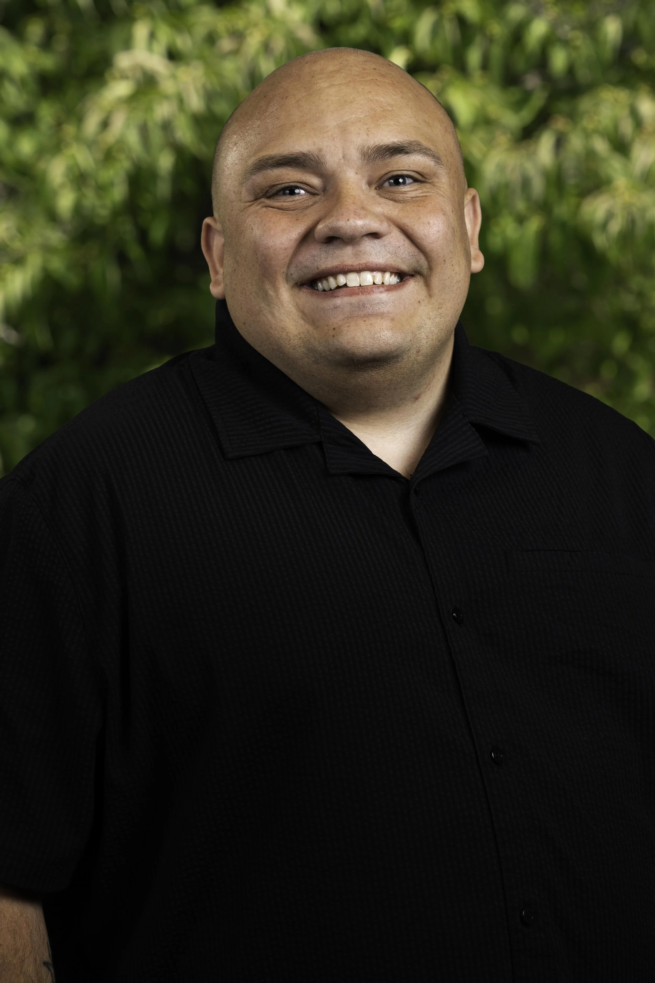 A smiling man wearing a camouflage cap with a red letter 'B', a black button-up shirt, and tattoos on both arms, holding a small snack in his hands.
