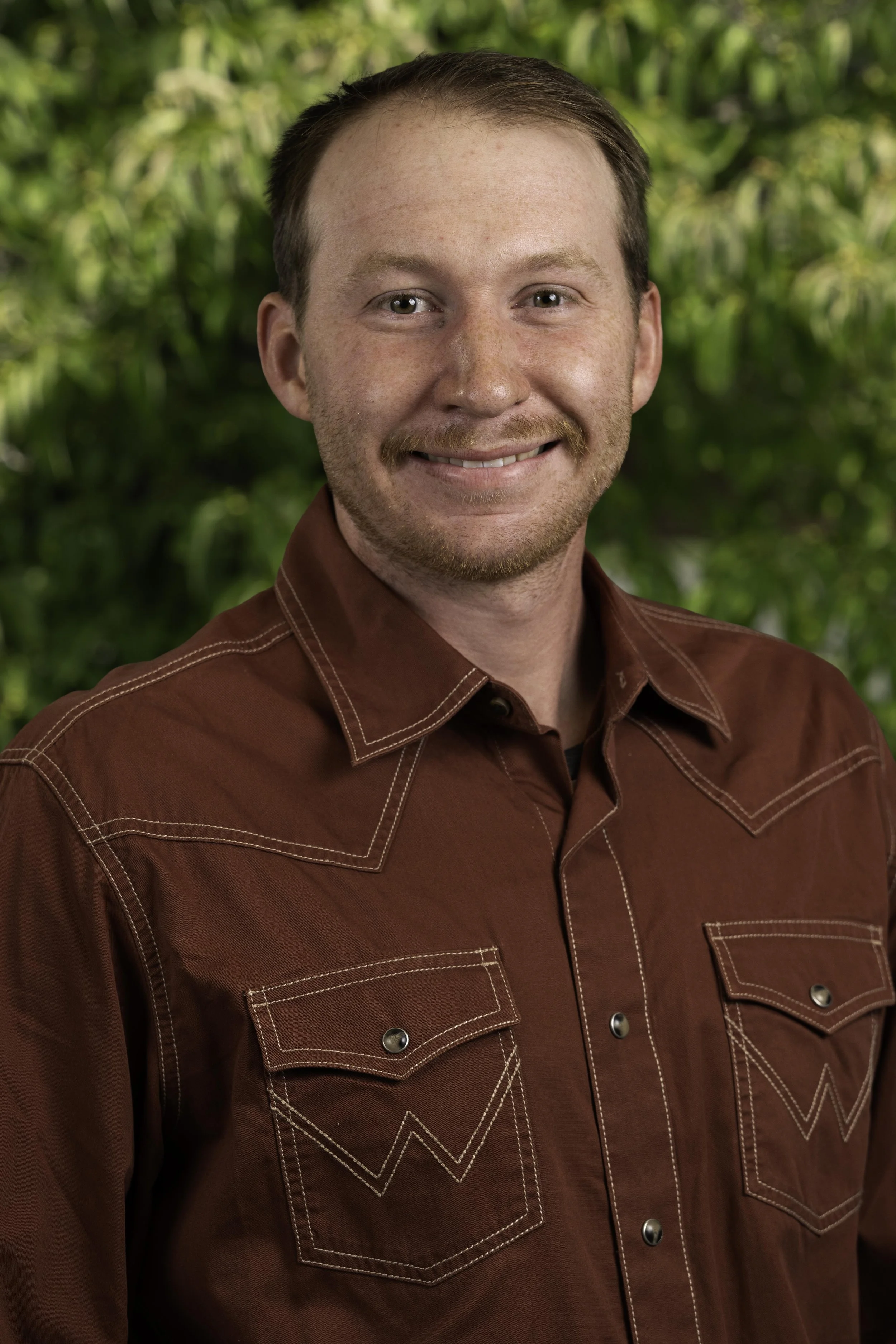 A man wearing a large white cowboy hat and a dark gray button-up shirt with colorful horizontal stripes smiling indoors.