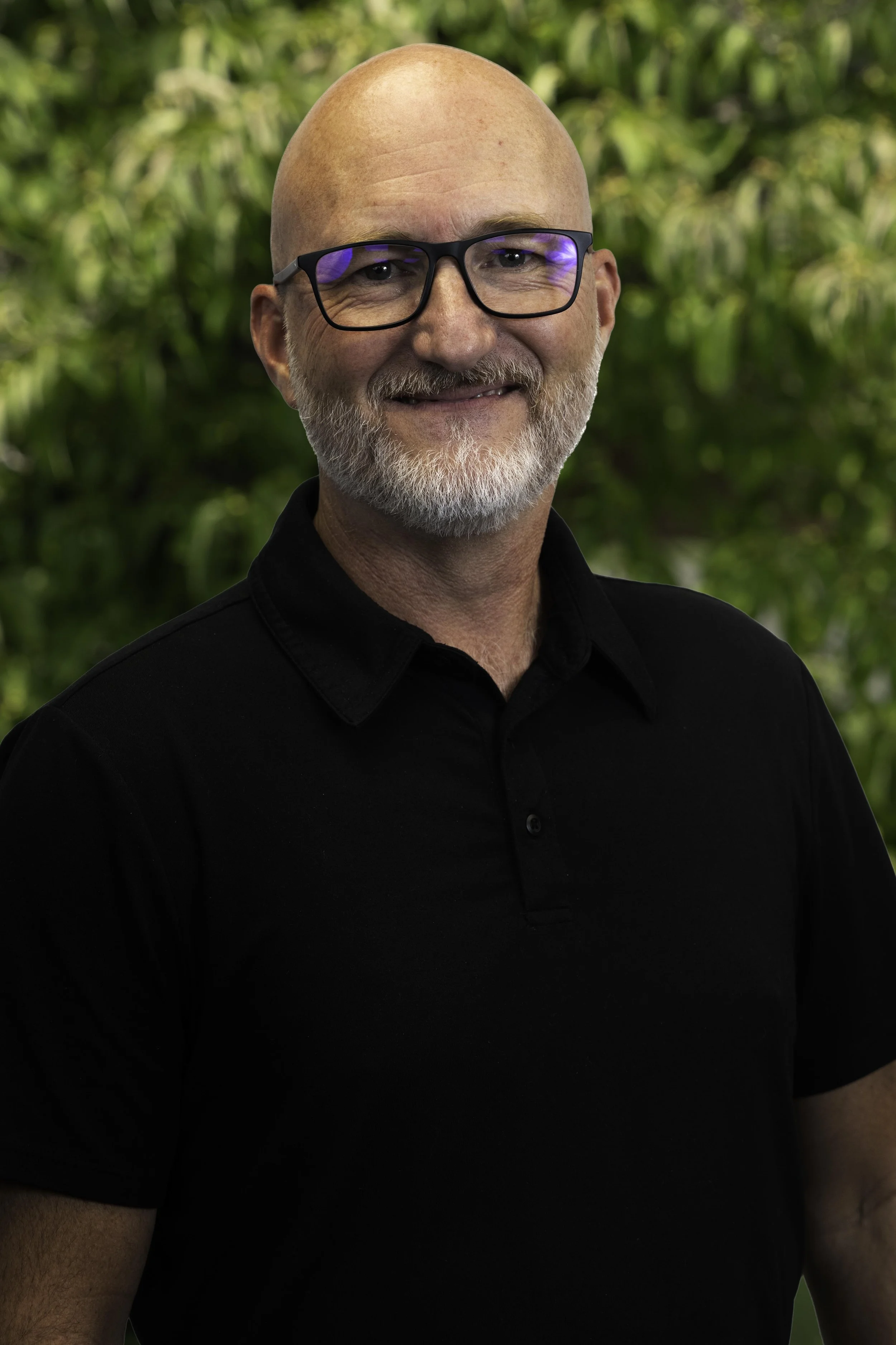 Portrait of a middle-aged man with a bald head, glasses, light beard, smiling, wearing a black shirt, standing outdoors with green foliage in the background.
