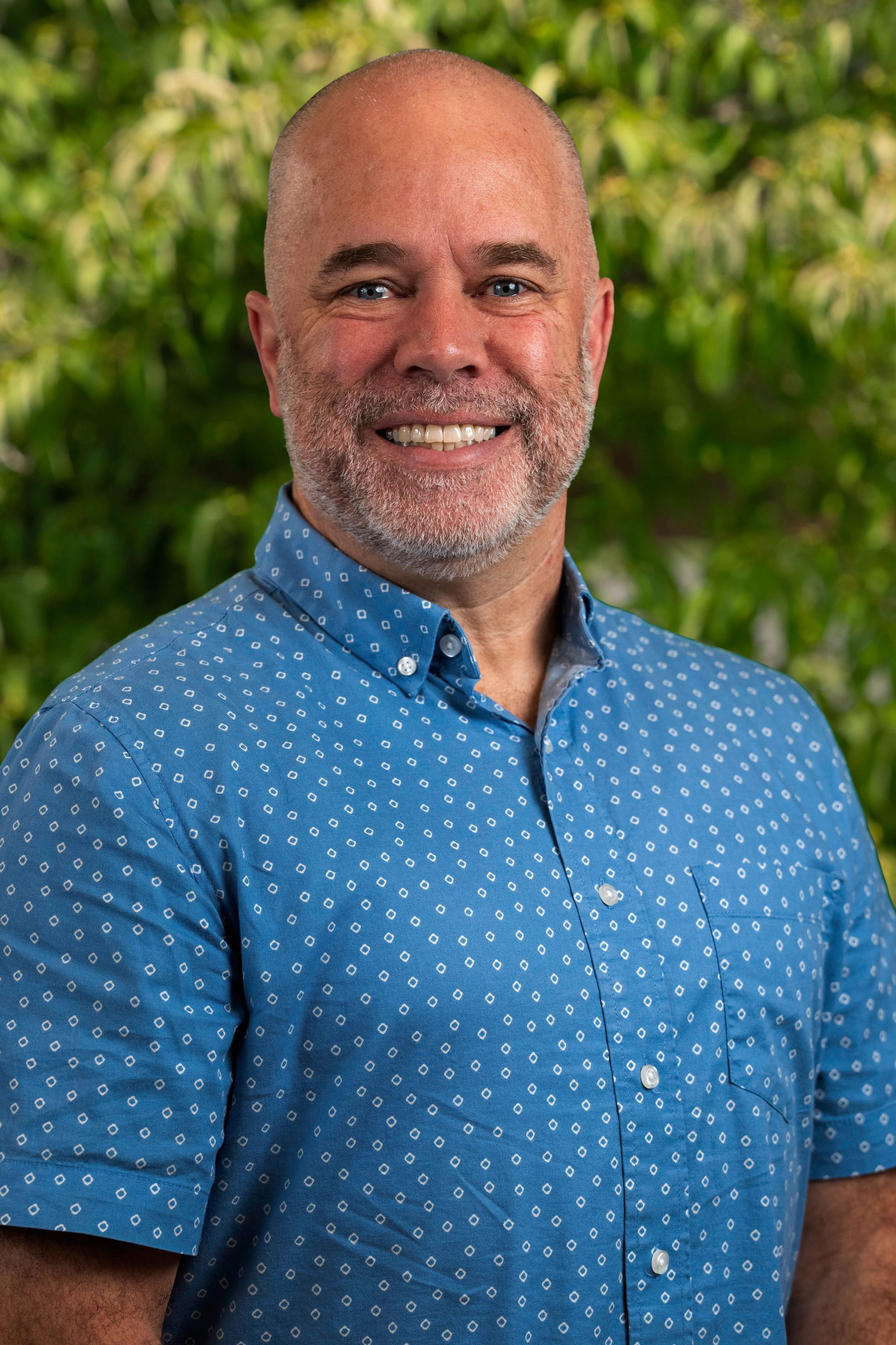 A smiling man with a beard and shaved head outdoors in a park on a sunny fall day, wearing a plaid shirt, with trees and a pathway in the background.