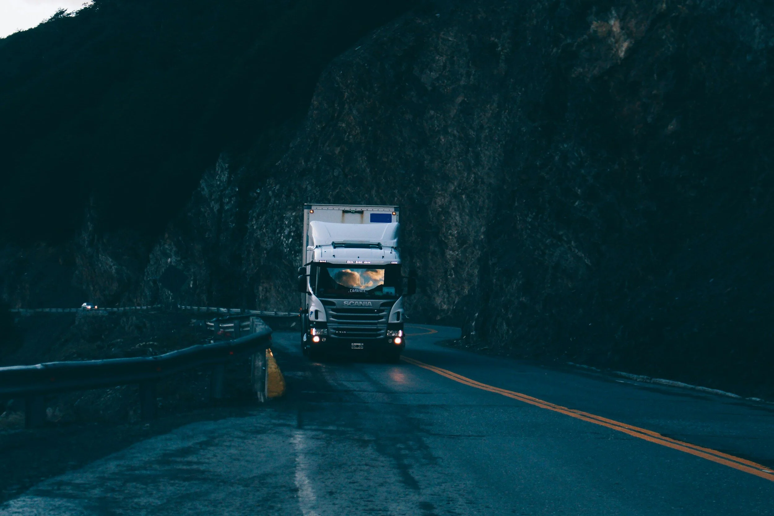 A white semi-truck driving along a highway through a mountainous area with rocky cliffs on both sides.