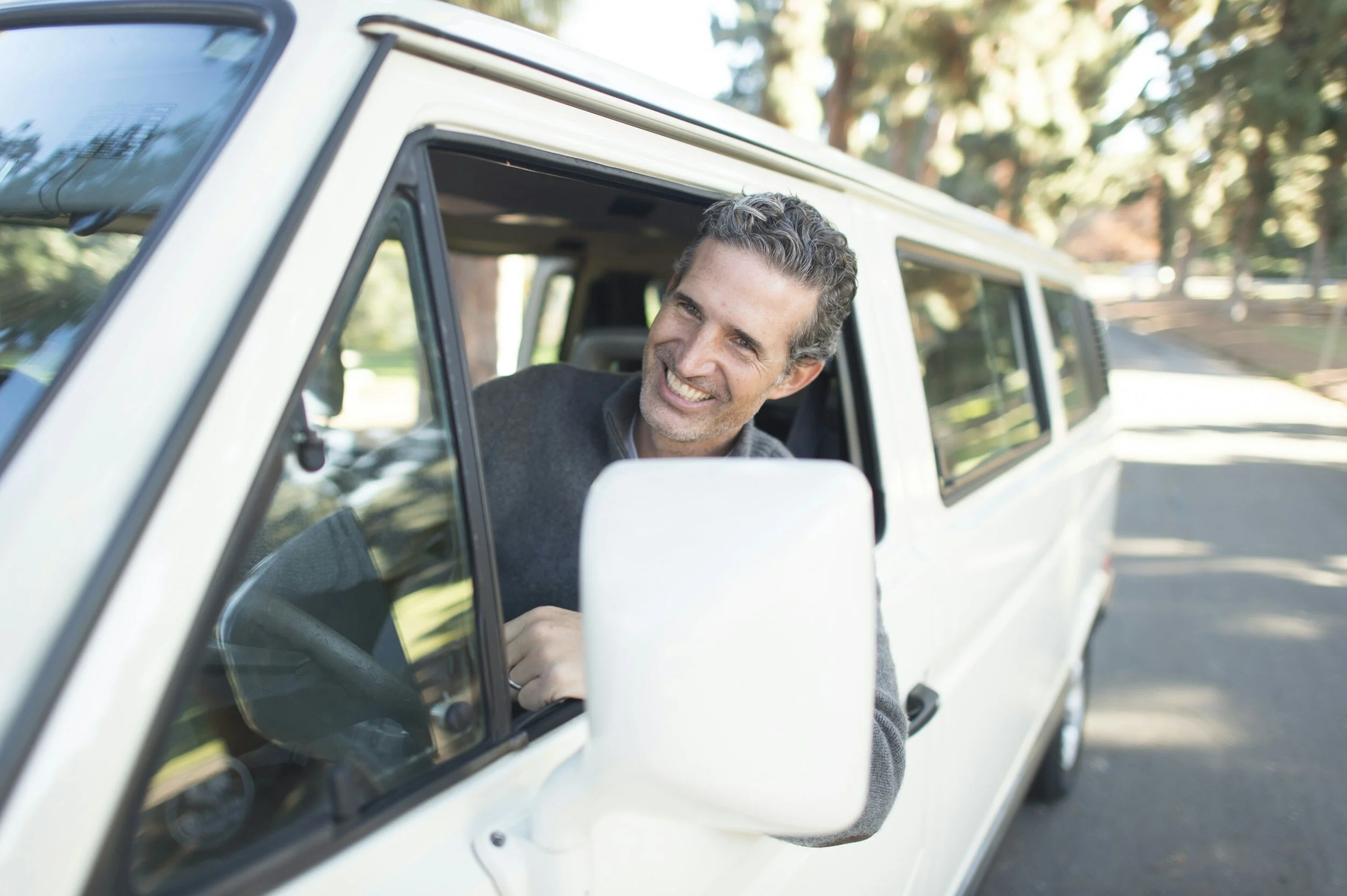 Smiling man leaning out of the driver's side window of a white vehicle parked on a tree-lined street.