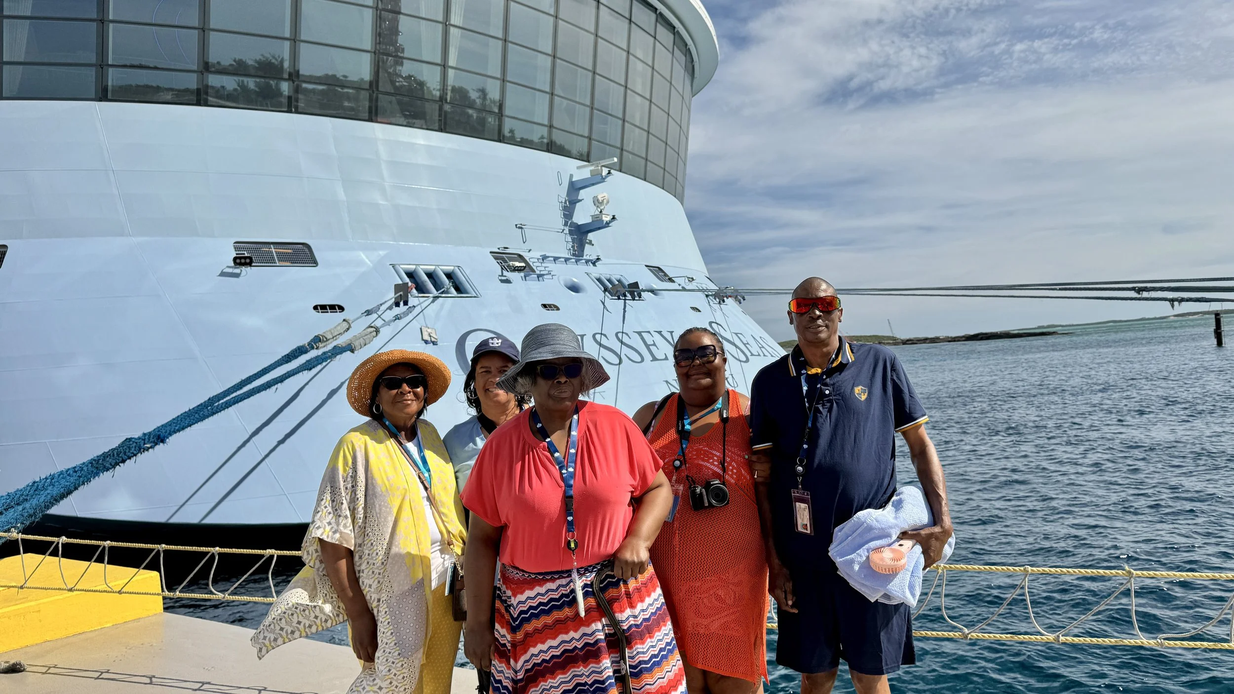 Group of five diverse adults standing in front of a large yacht at a dock, with water and a cloudy sky in the background.