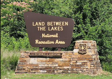 Sign reading 'Land Between the Lakes, National Recreation Area, U.S. Department of Agriculture' with trees and greenery in the background.