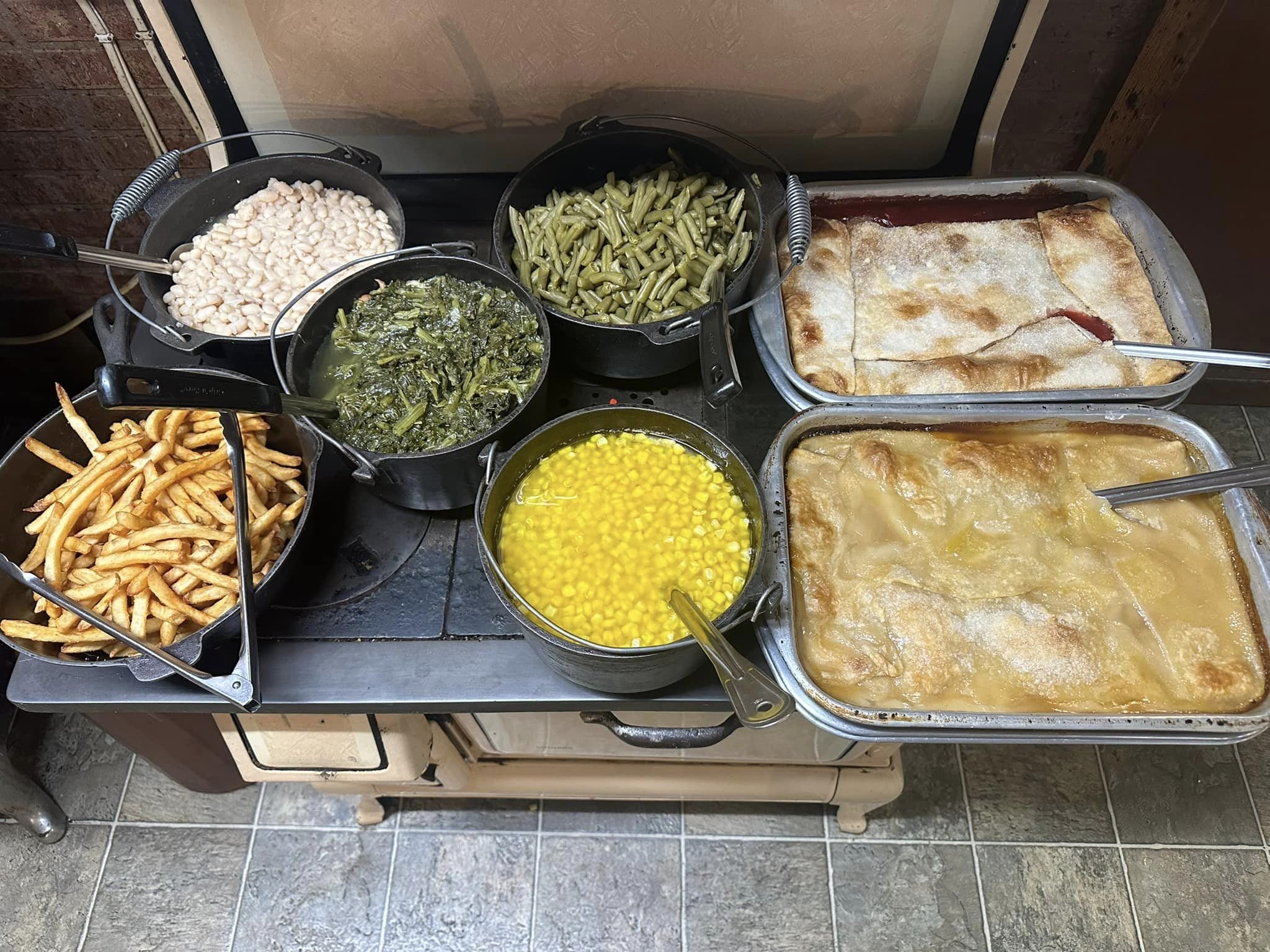 A collection of different cooked foods on a counter, including fried potatoes, white beans, green beans, asparagus, corn, and two trays of baked dish, possibly lasagna or casserole.