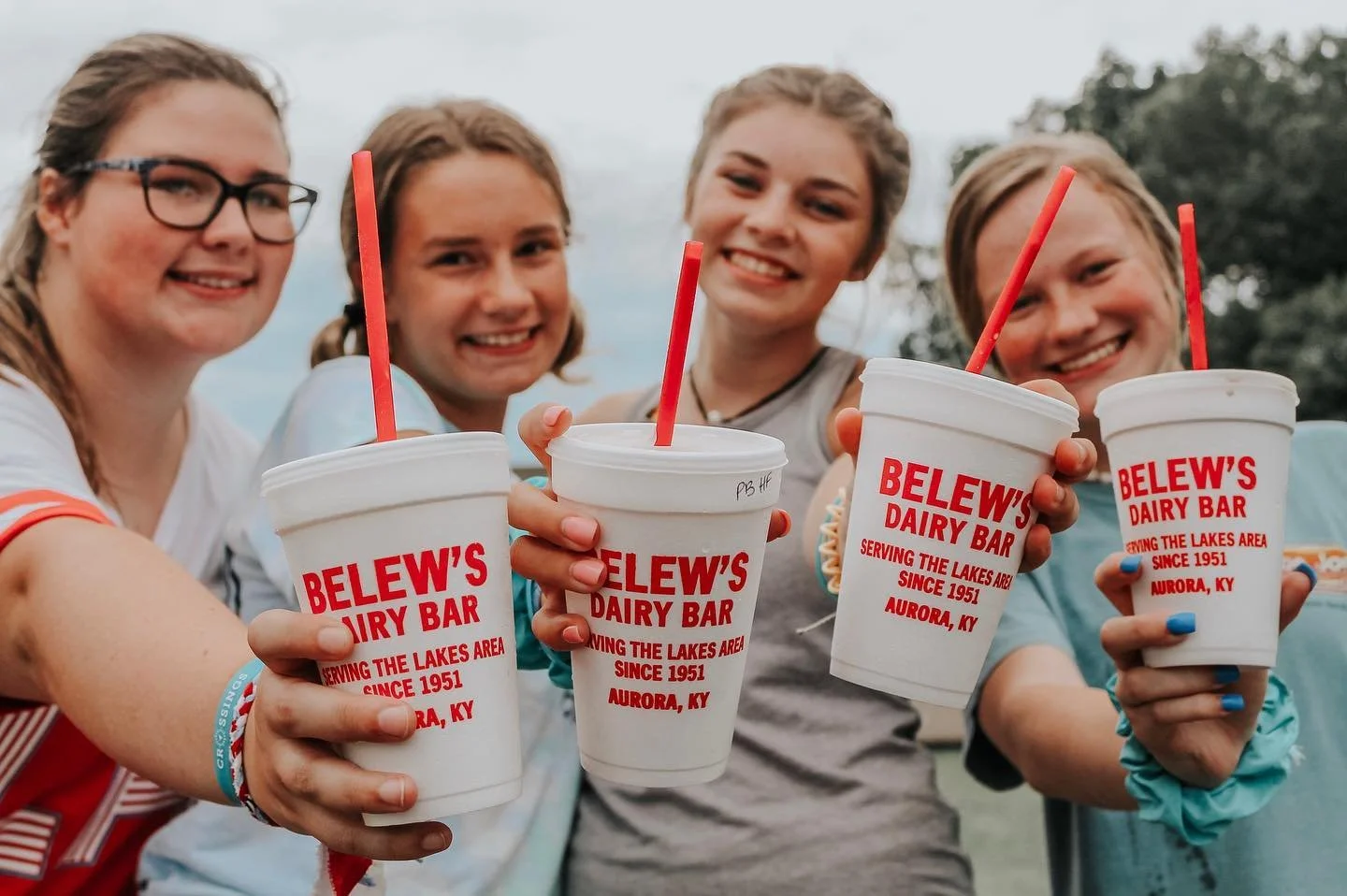 Four young women outdoors smiling and holding large cups of Dairy Bar milkshakes, with red straws, branded with 'Belew's Dairy Bar' from Aurora, Kentucky.