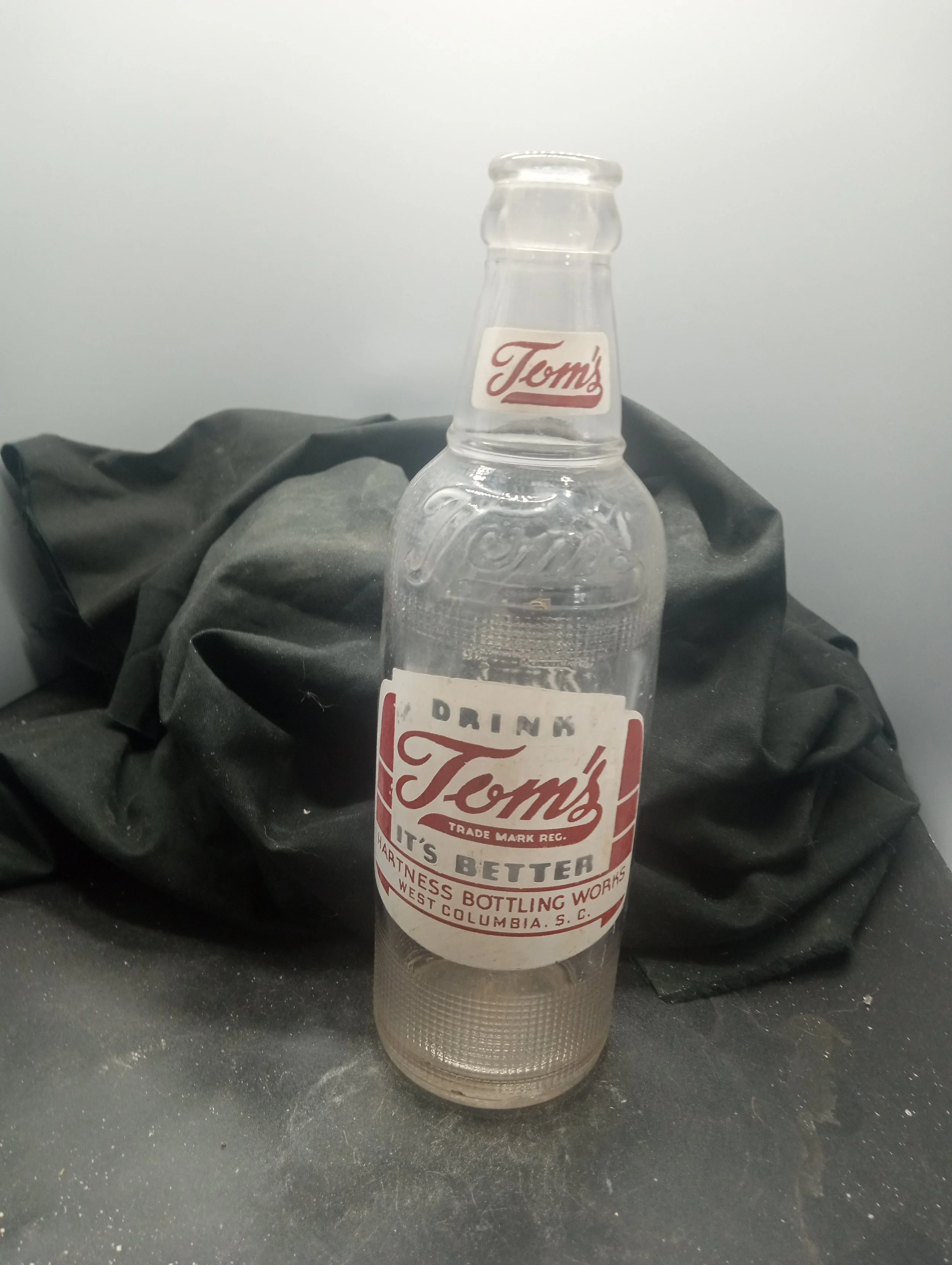 AGE - 1954 - TYPE - MACHINE - FRONT - (neck) TOM'S (embossed at shoulder) TOM'S (body) DRINK  / TOM'S / TRADE MARK REG. / IT'S BETTER / HARTNESS BOTTLING WORKS / WEST COLUMBIA, S.C. - BACK - TASTE THE / DIFFERENCE / TOM'S / IS / BETTER / CONTENTS 12 