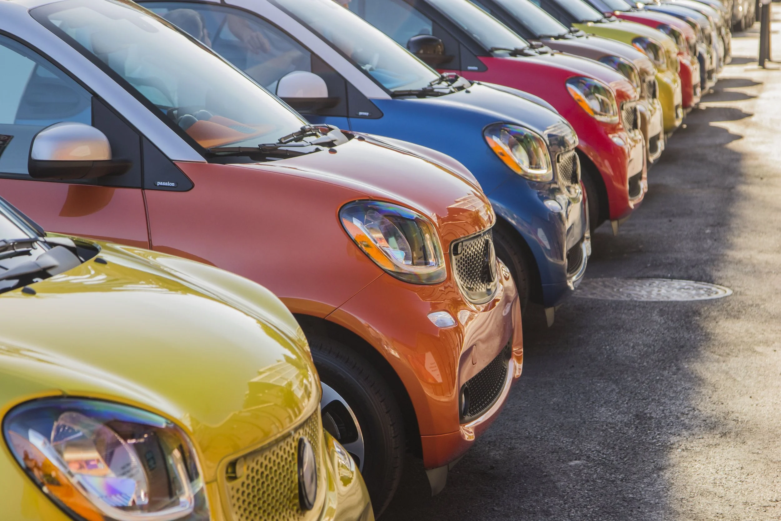Multiple compact cars in yellow, orange, blue, red, and black, parked in a row at a dealership or parking lot, viewed from the front and side.