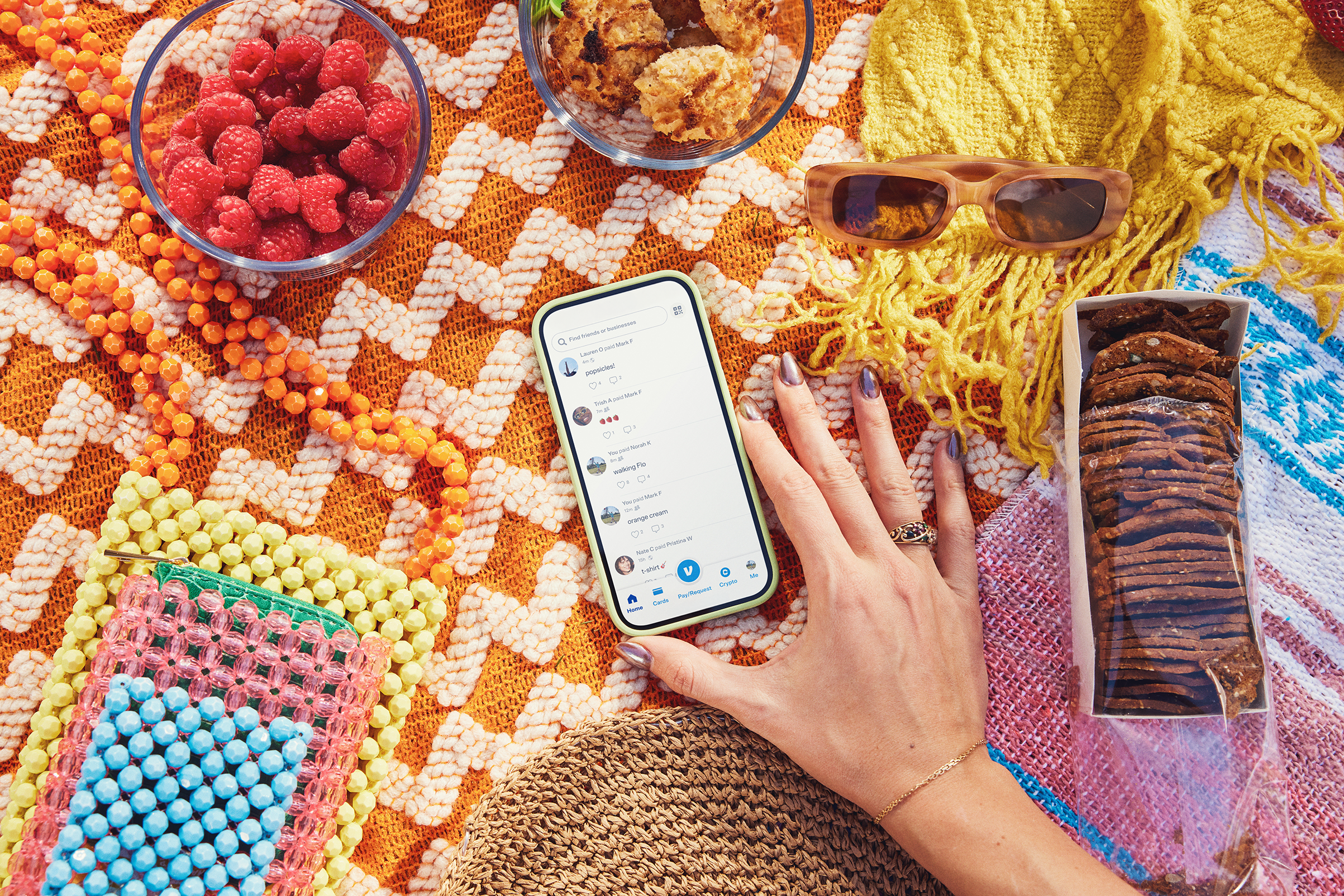 Colorful beaded jewelry, raspberries, cookies, sunglasses, a smartphone showing a social media feed, and a box of cookies on a vibrant, patterned textile background.