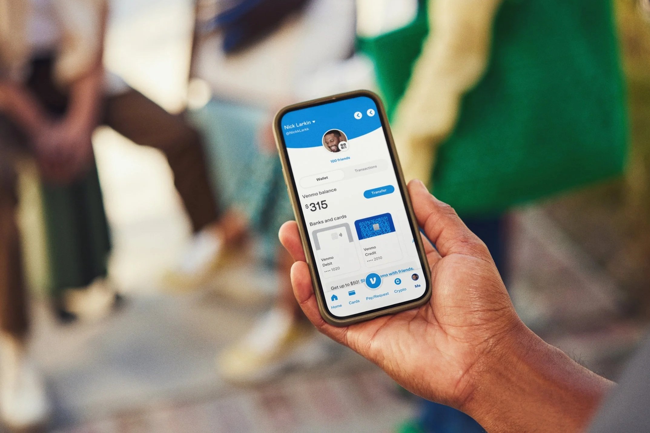 Person holding a smartphone displaying a Venmo profile with a balance of $315, in a blurred background of people and colorful surroundings.
