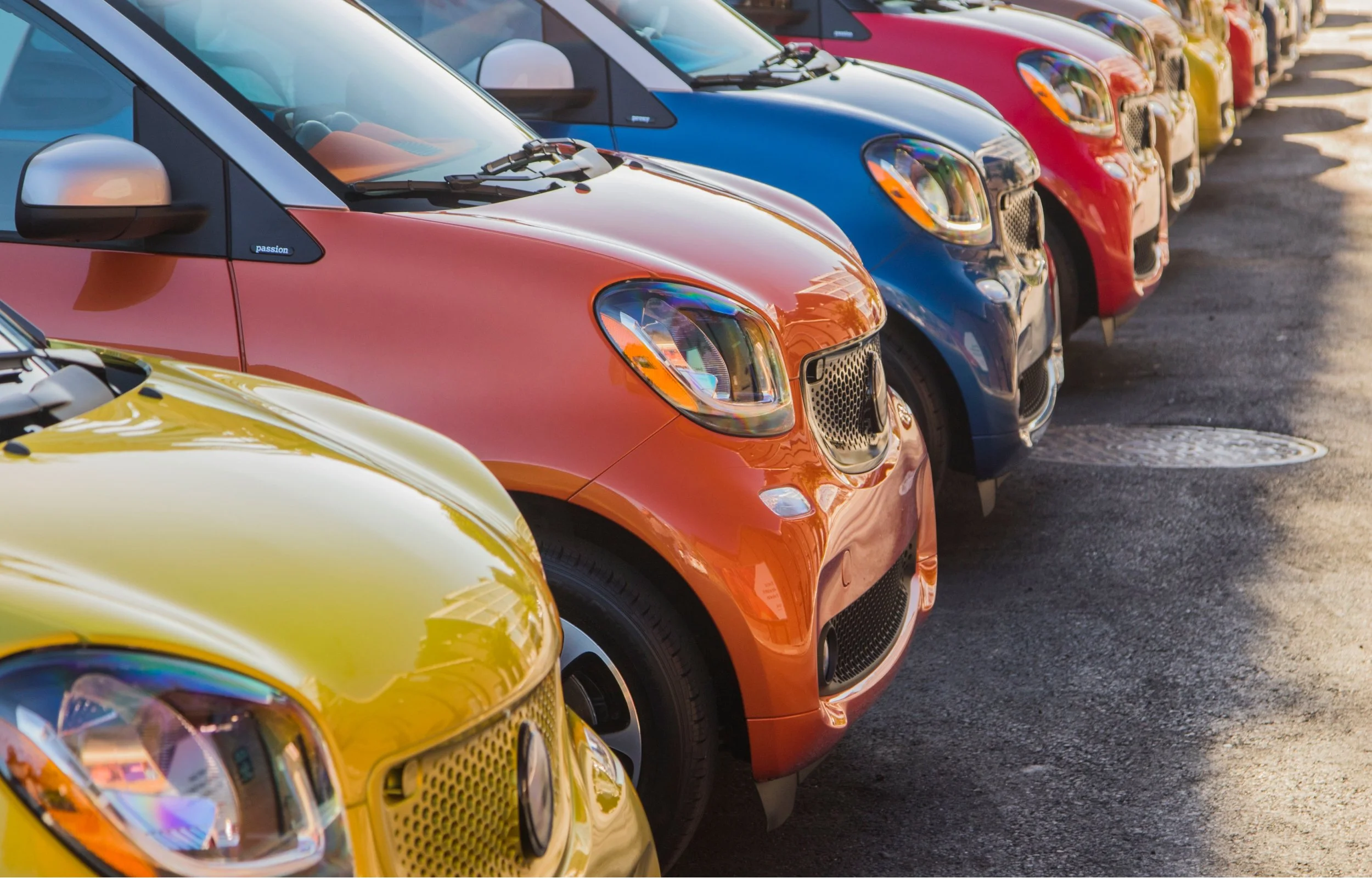 Lineup of colorful compact cars parked on a street during daytime, including a yellow, orange, blue, and red vehicle, with reflections visible on their shiny surfaces.