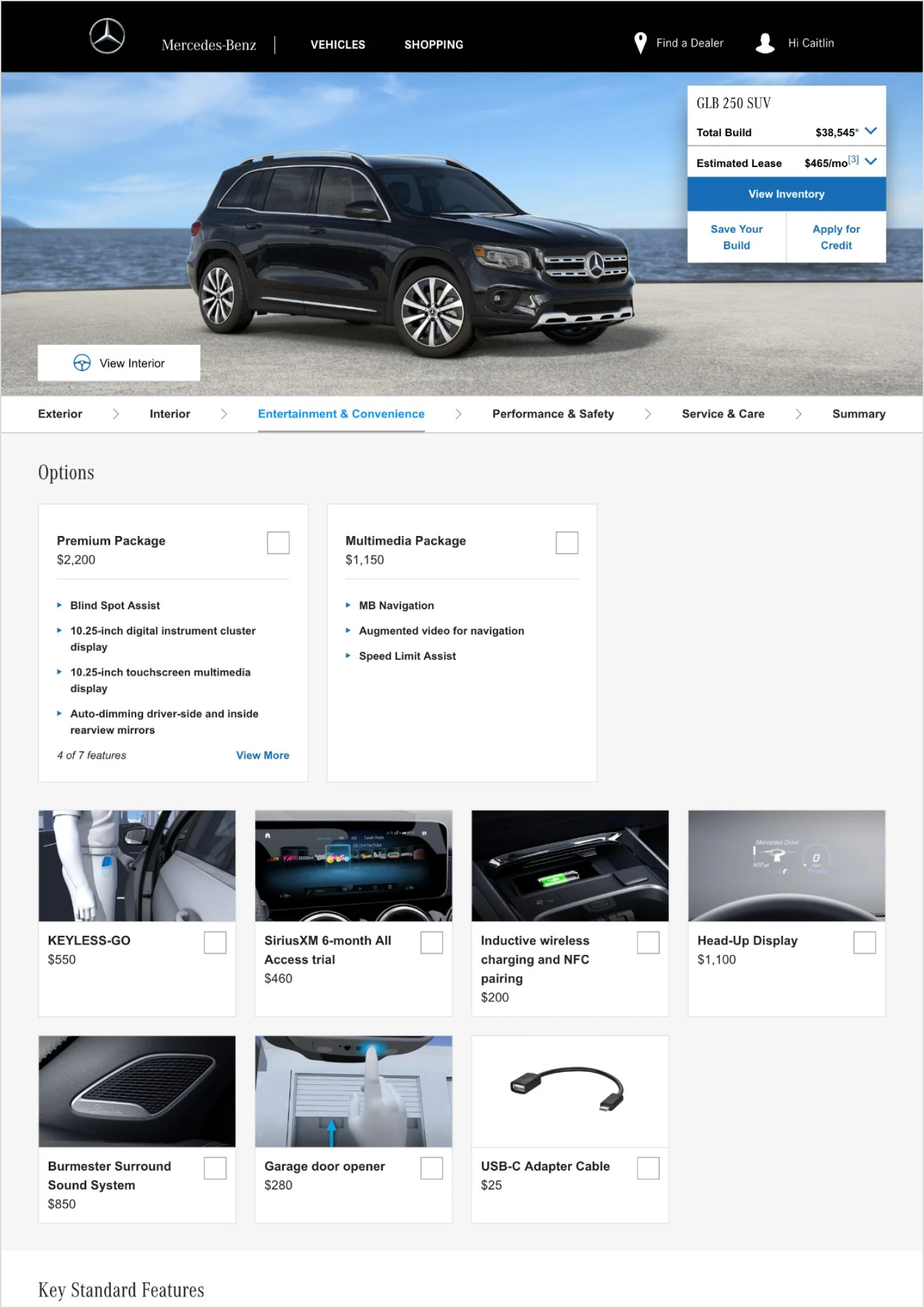 Screenshot of a Mercedes-Benz vehicle customization webpage featuring a black SUV by the ocean. The menu includes options for exterior, interior, entertainment and convenience, performance and safety, service and care, and a summary. Available options include packages like premium, multimedia, keyless-go, SiriusXM trial, wireless charging, head-up display, and accessories like a surround sound system, garage door opener, and USB-C adapter cable.