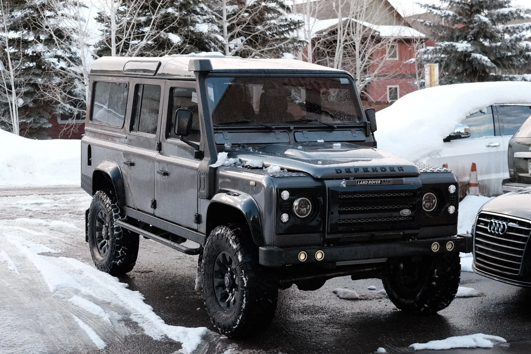 A black Land Rover Defender SUV parked on a snow-covered road, with snow on its hood and windshield, and a white car parked beside it. Snow-covered trees and buildings are in the background.