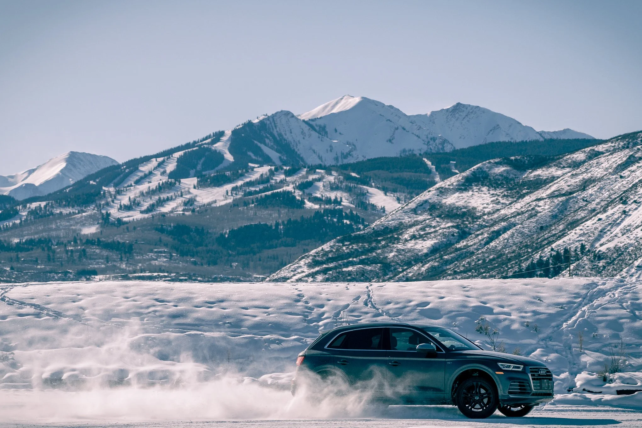 Black Audi SUV driving on snowy mountain road with snow-covered mountains in the background.
