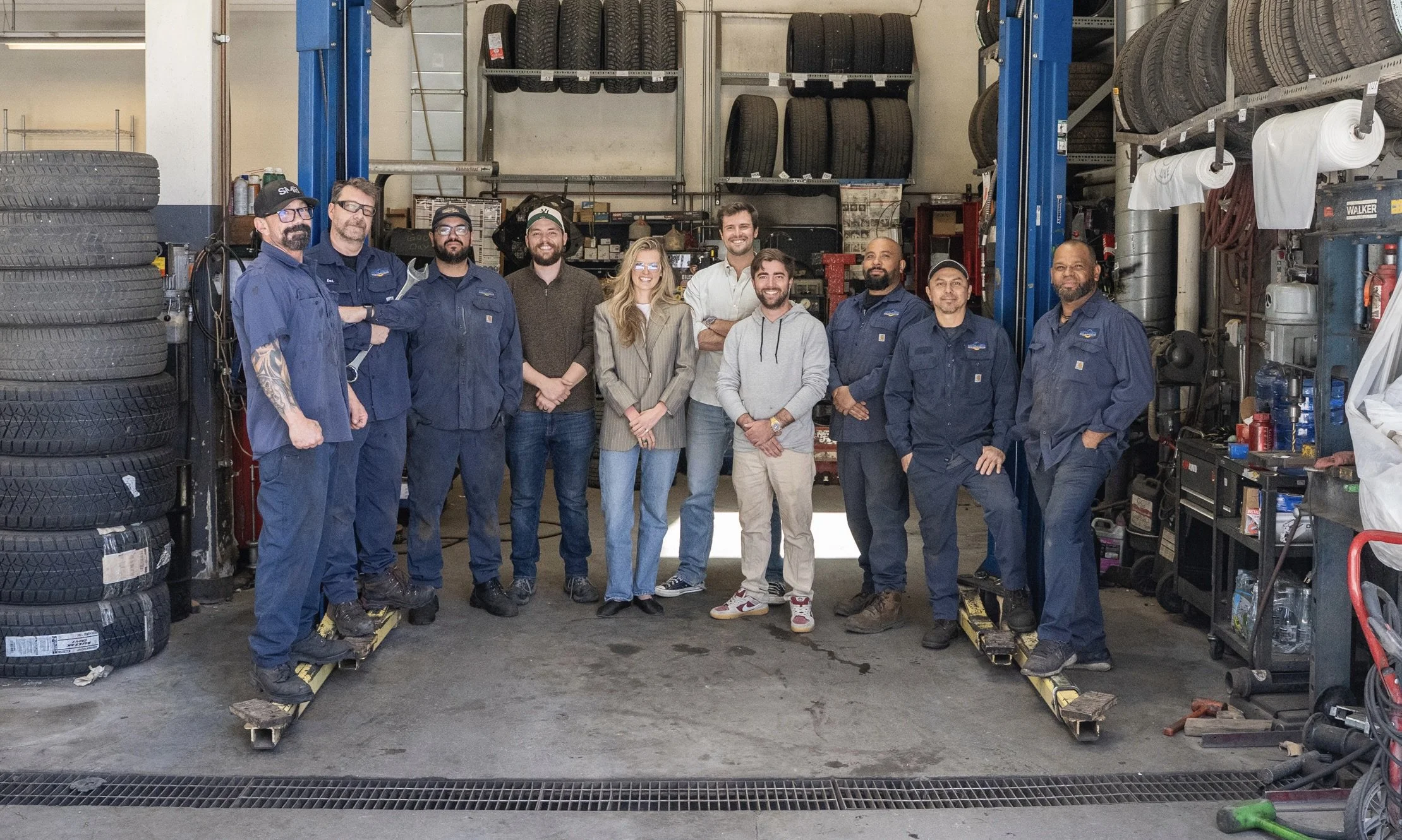 Group of eleven people, including mechanical and office staff, standing together inside an auto repair shop, with tires and auto parts visible in the background.