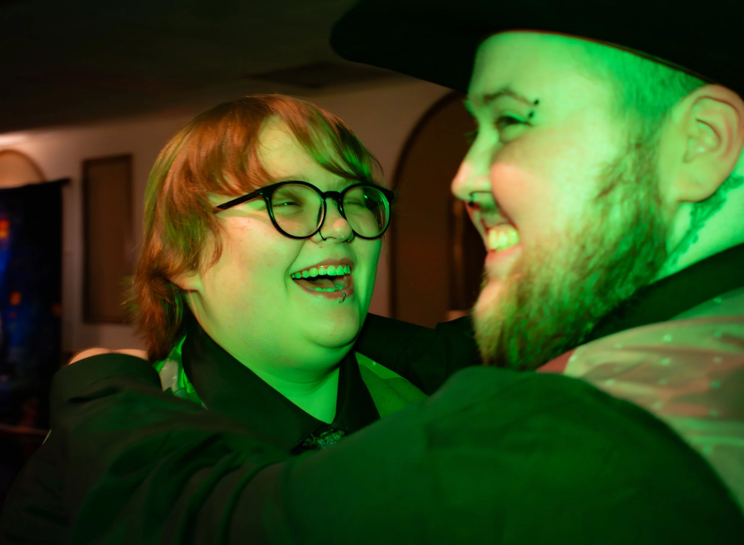 Two people, one with glasses and the other wearing a hat, are smiling and looking at each other in a dimly lit room with green lighting.