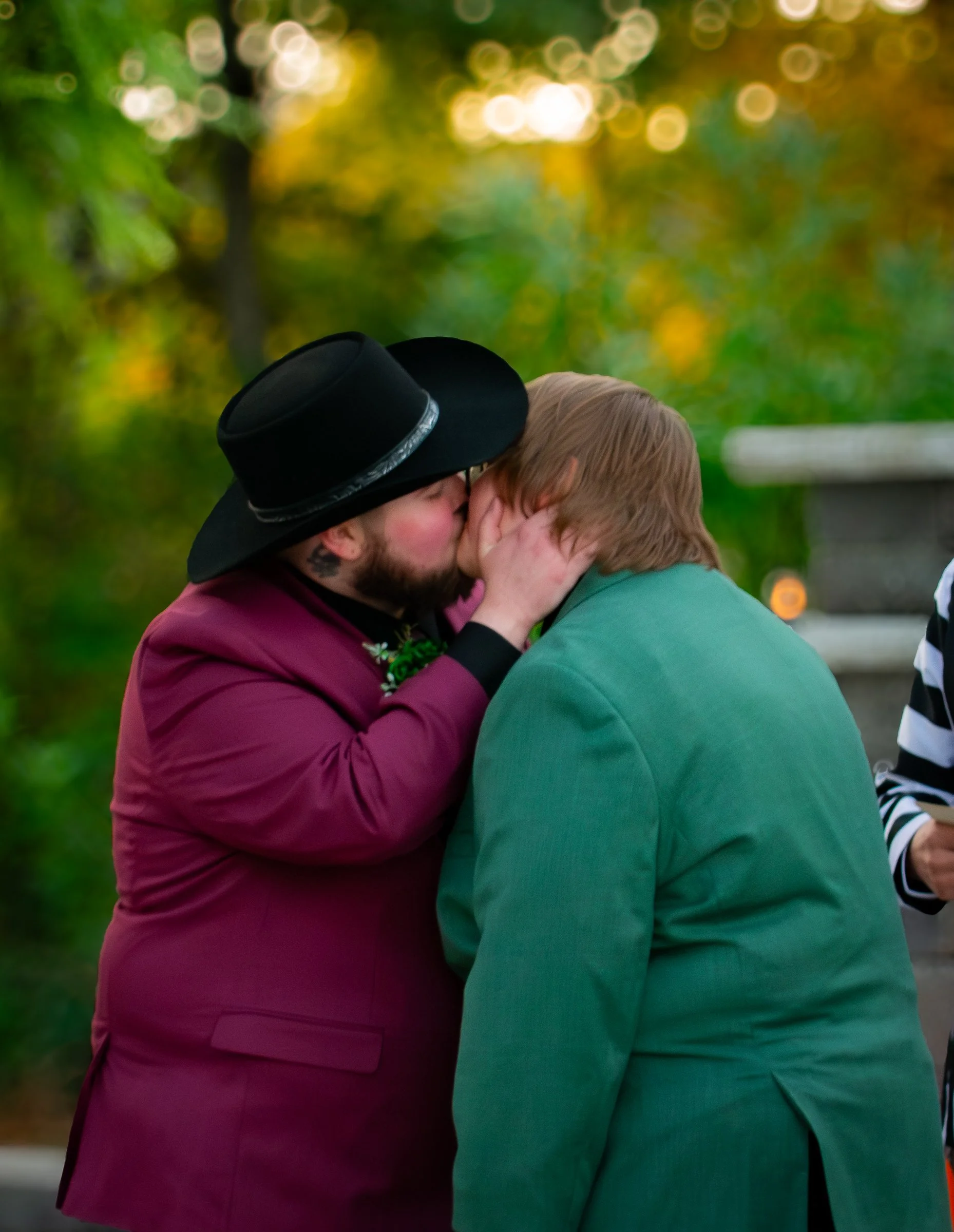 Two men, one wearing a maroon jacket and black hat, the other in a green coat, are sharing a kiss on the forehead outdoors with blurred autumn trees in the background.