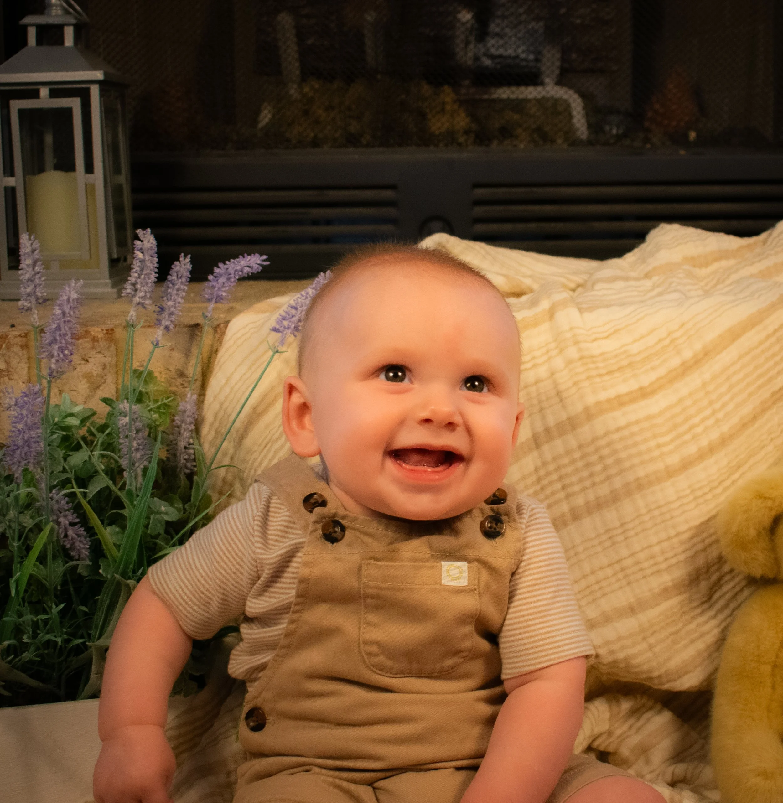 Close-up of a smiling baby wearing a beige outfit with black buttons, sitting on a beige blanket in front of a fireplace with a fire. There are lavender flowers in front of the baby, and a lantern with a candle is on the side. The fireplace has a screen and decorative items on top.