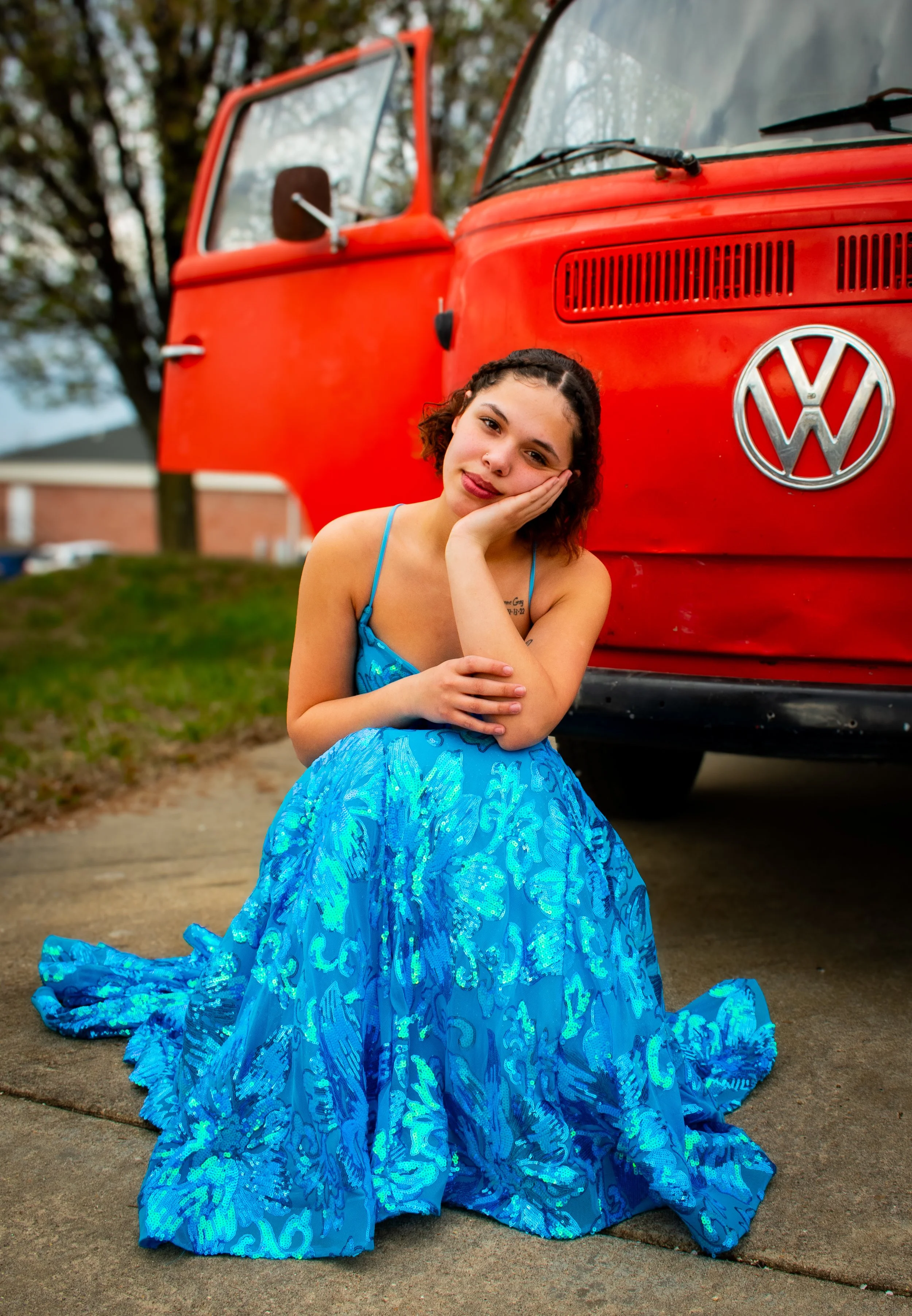 A young woman in a blue dress with a flowing, patterned skirt is sitting on a sidewalk, resting her face on her hand. She is in front of a red vintage Volkswagen van parked outdoors with trees in the background.