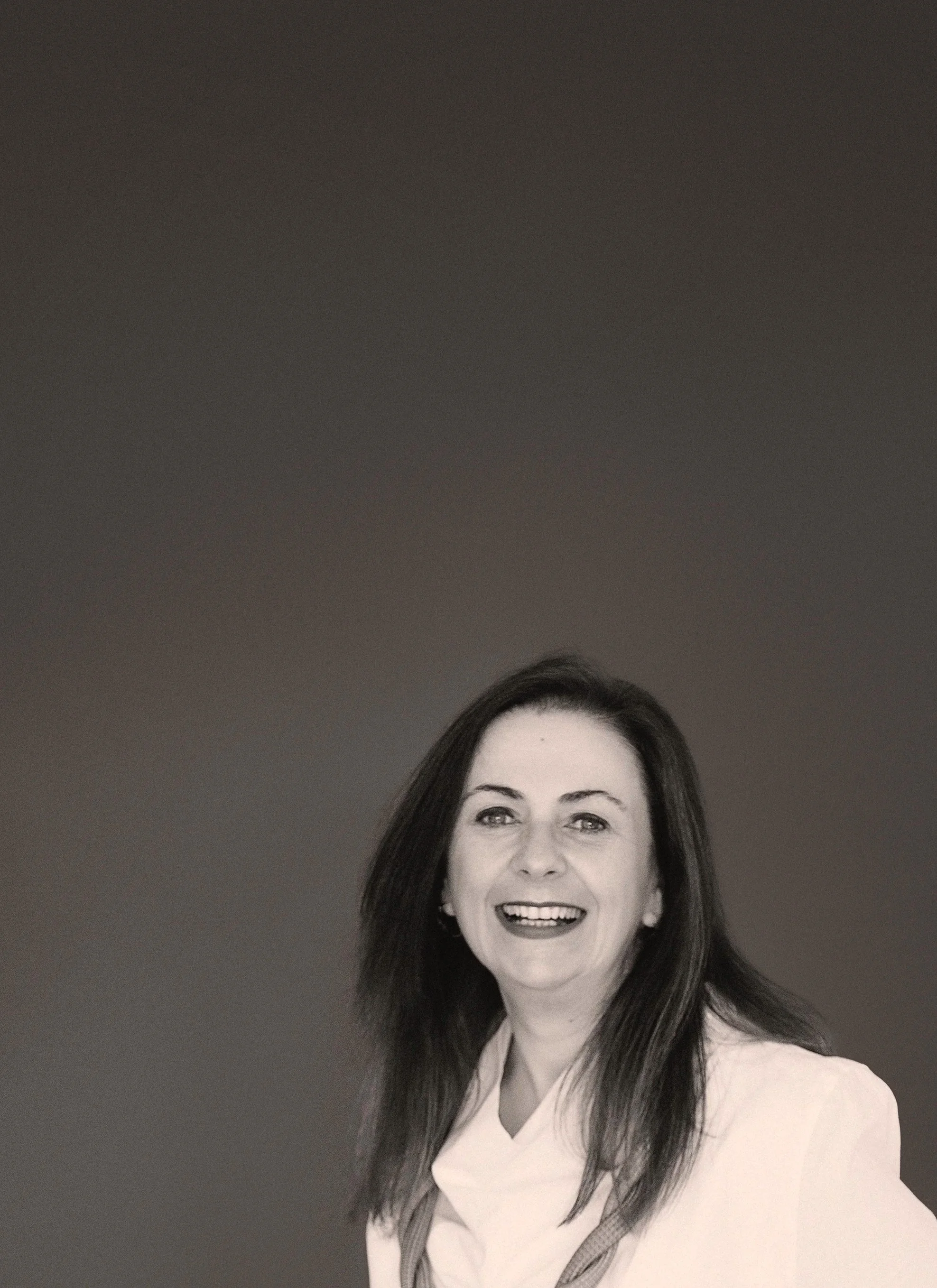 Black-and-white portrait of a smiling woman with long dark hair, wearing a light-colored blouse and jacket, against a plain background.