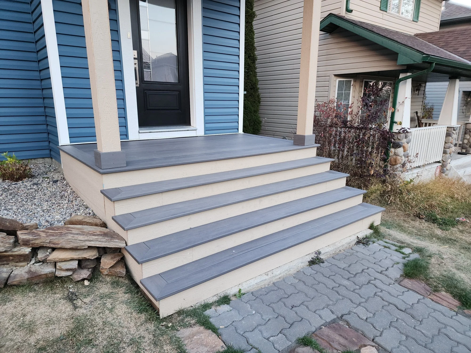 Front porch with gray steps, beige and blue siding, green gutter, and plants in the garden.